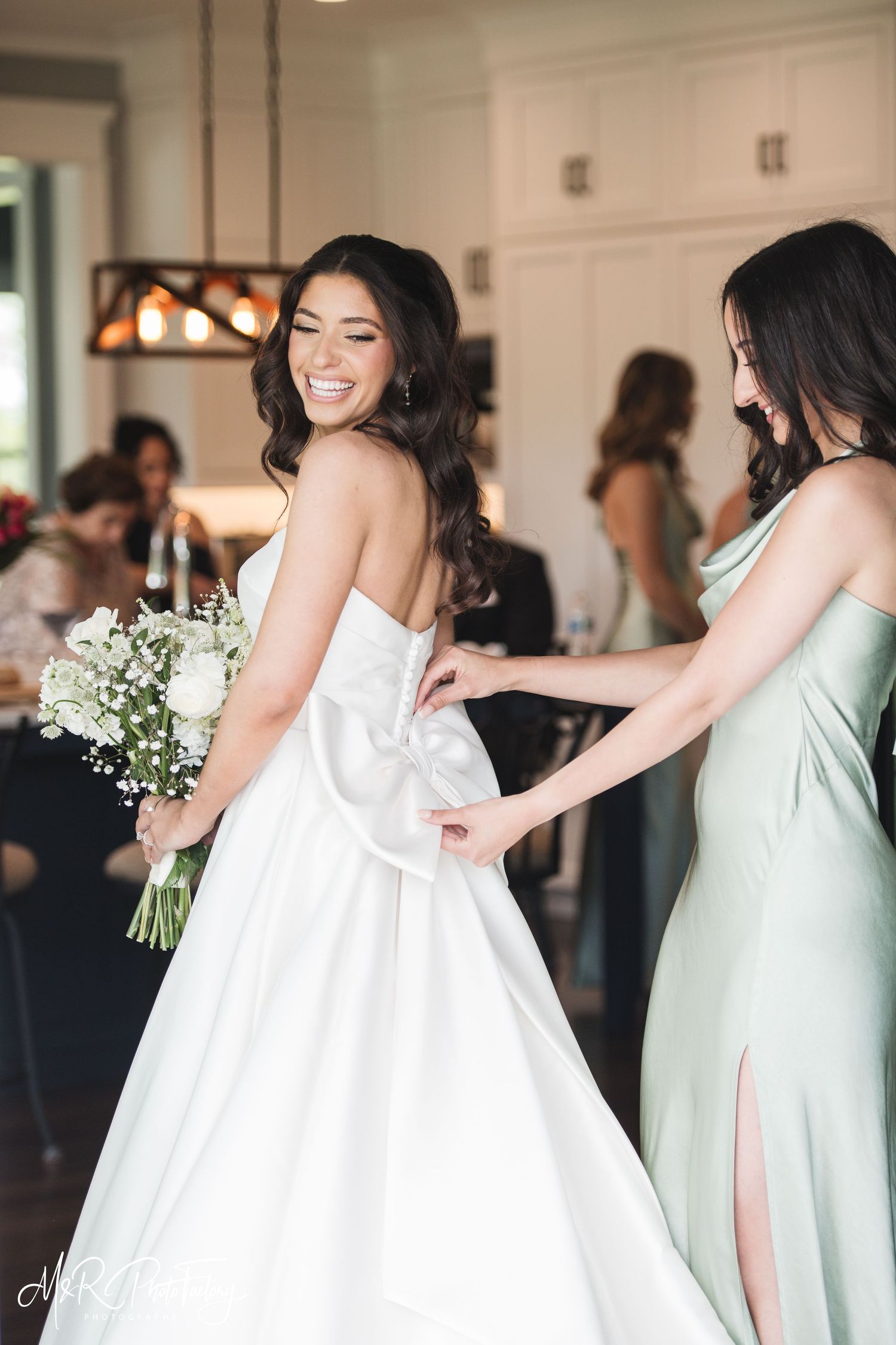 Radiant bride holding white floral bouquet while bridesmaid adjusts bow on back of her strapless wedding dress.