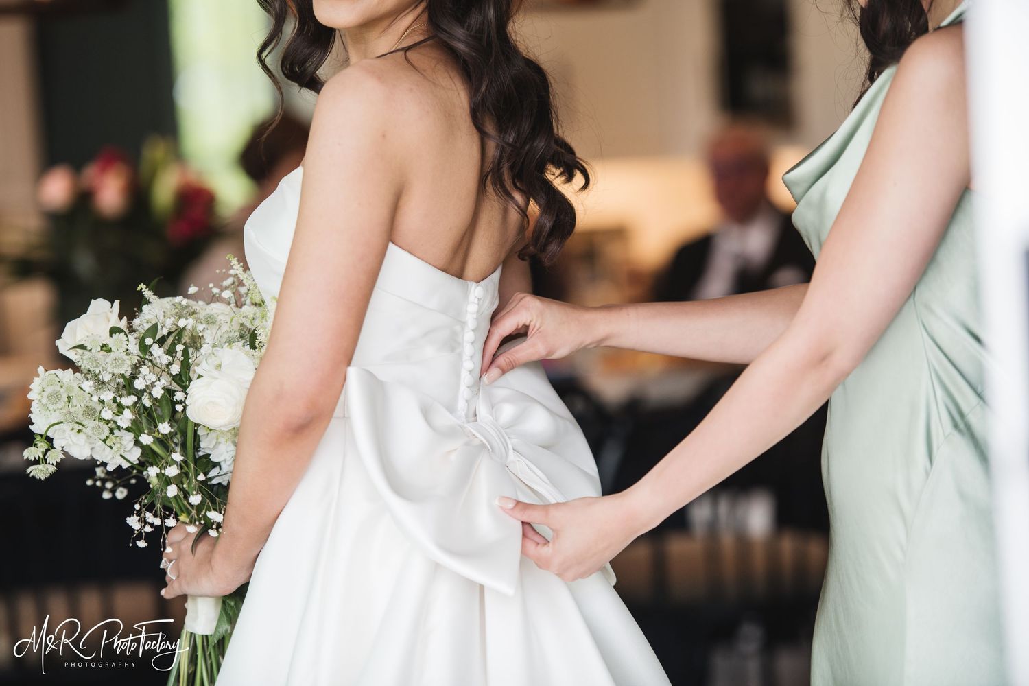 Close-up of bridesmaid tying large satin bow on back of bride's classic white strapless wedding gown.
