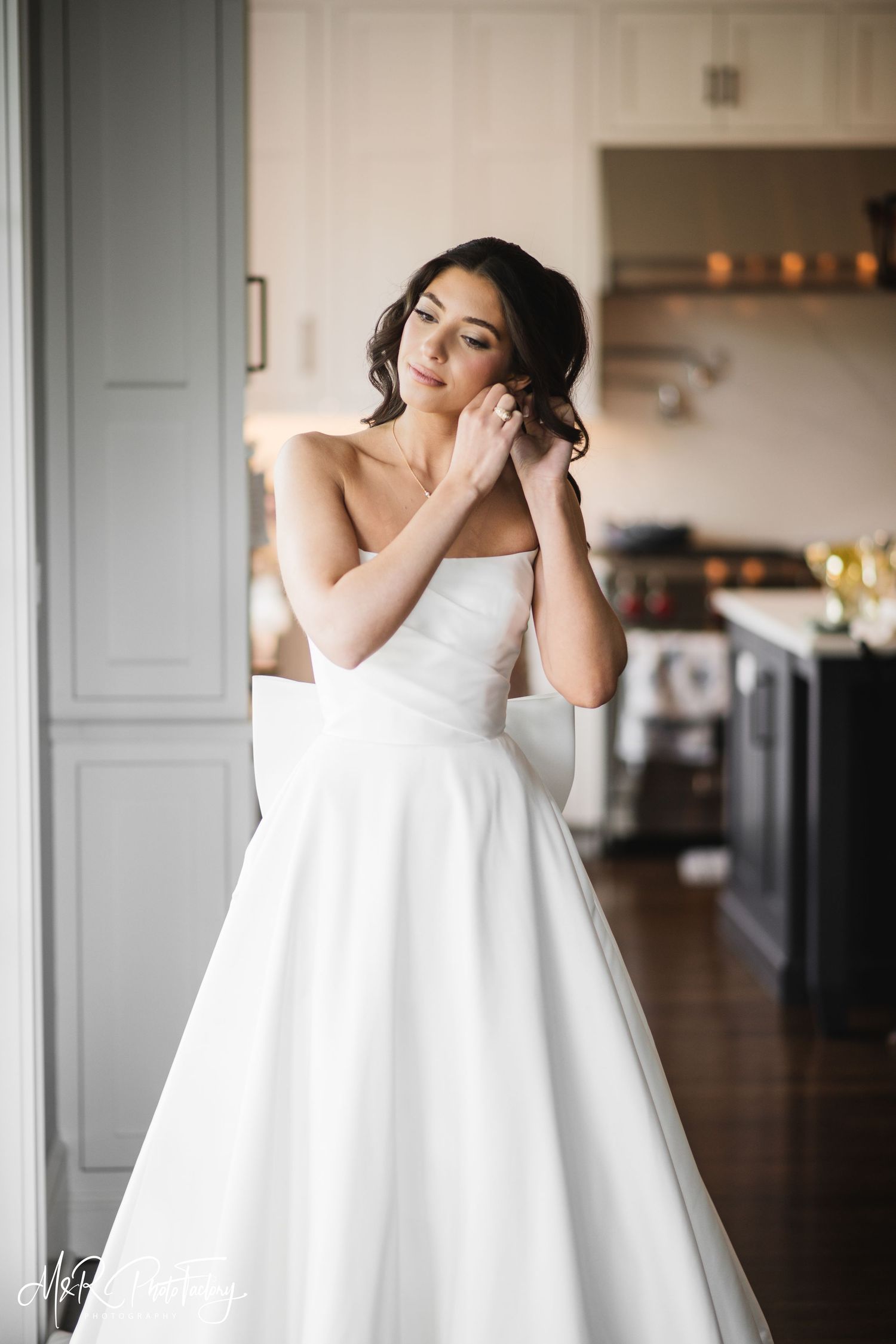 Elegant bride in strapless white ball gown adjusting earring while standing in a bright kitchen setting.