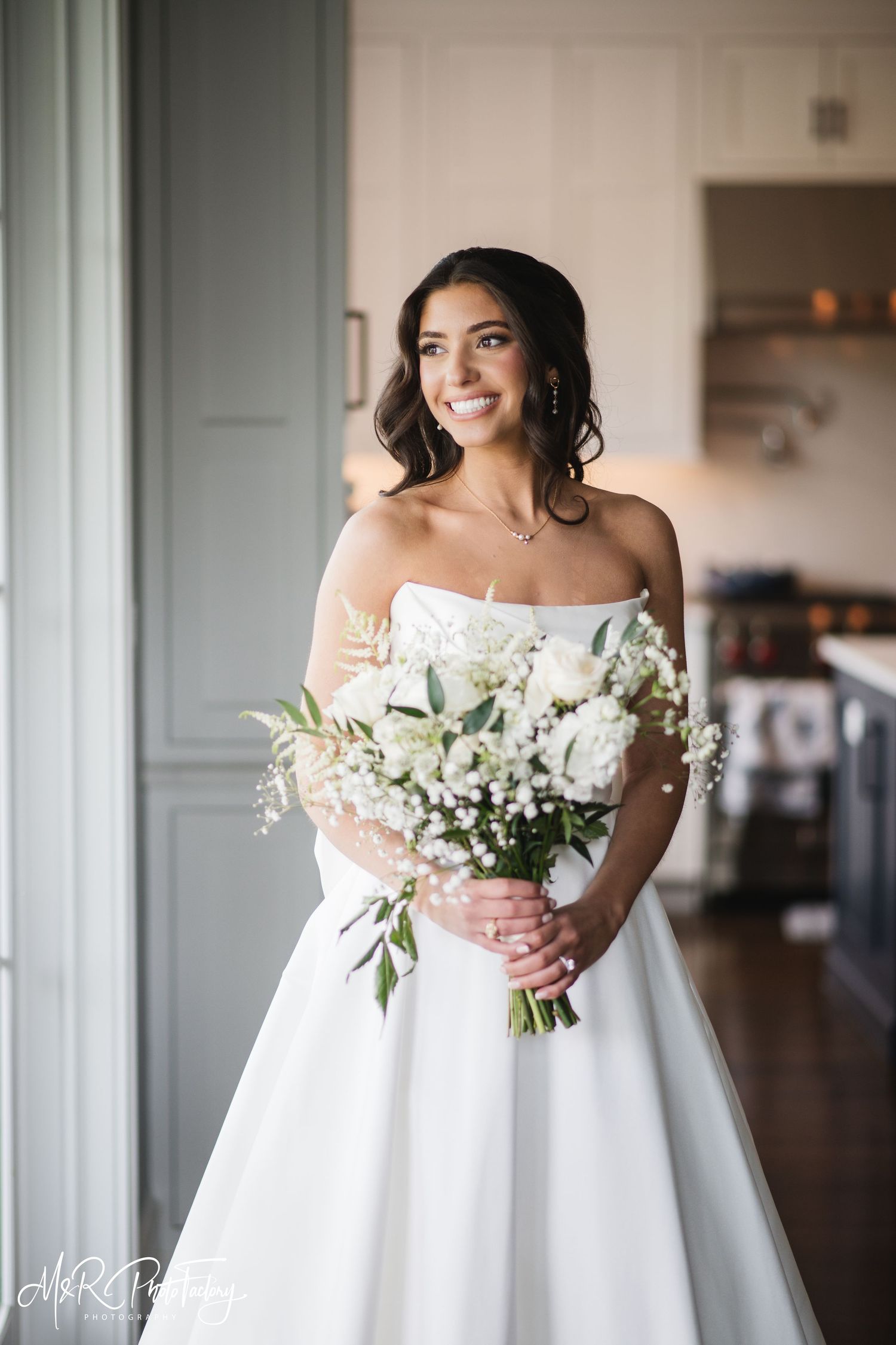 Beautiful bride in strapless white gown smiling while holding lush white floral bouquet with greenery.