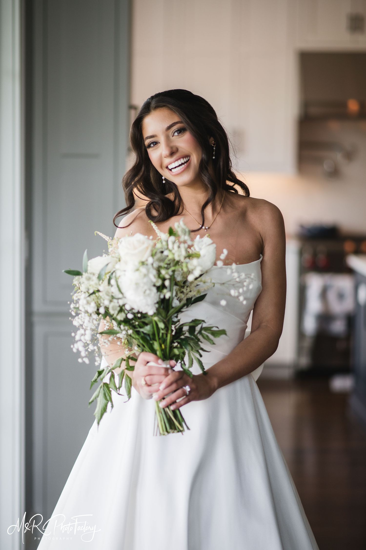Glowing bride in strapless white wedding gown laughing while holding elegant white floral bridal bouquet.