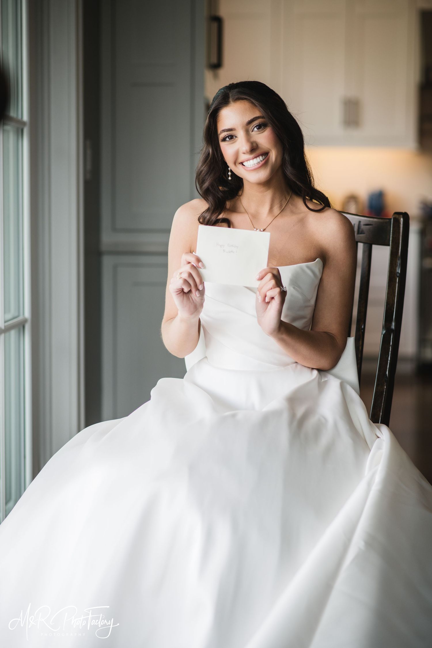 Smiling bride in strapless white gown sitting in chair holding a wedding letter card from her groom.