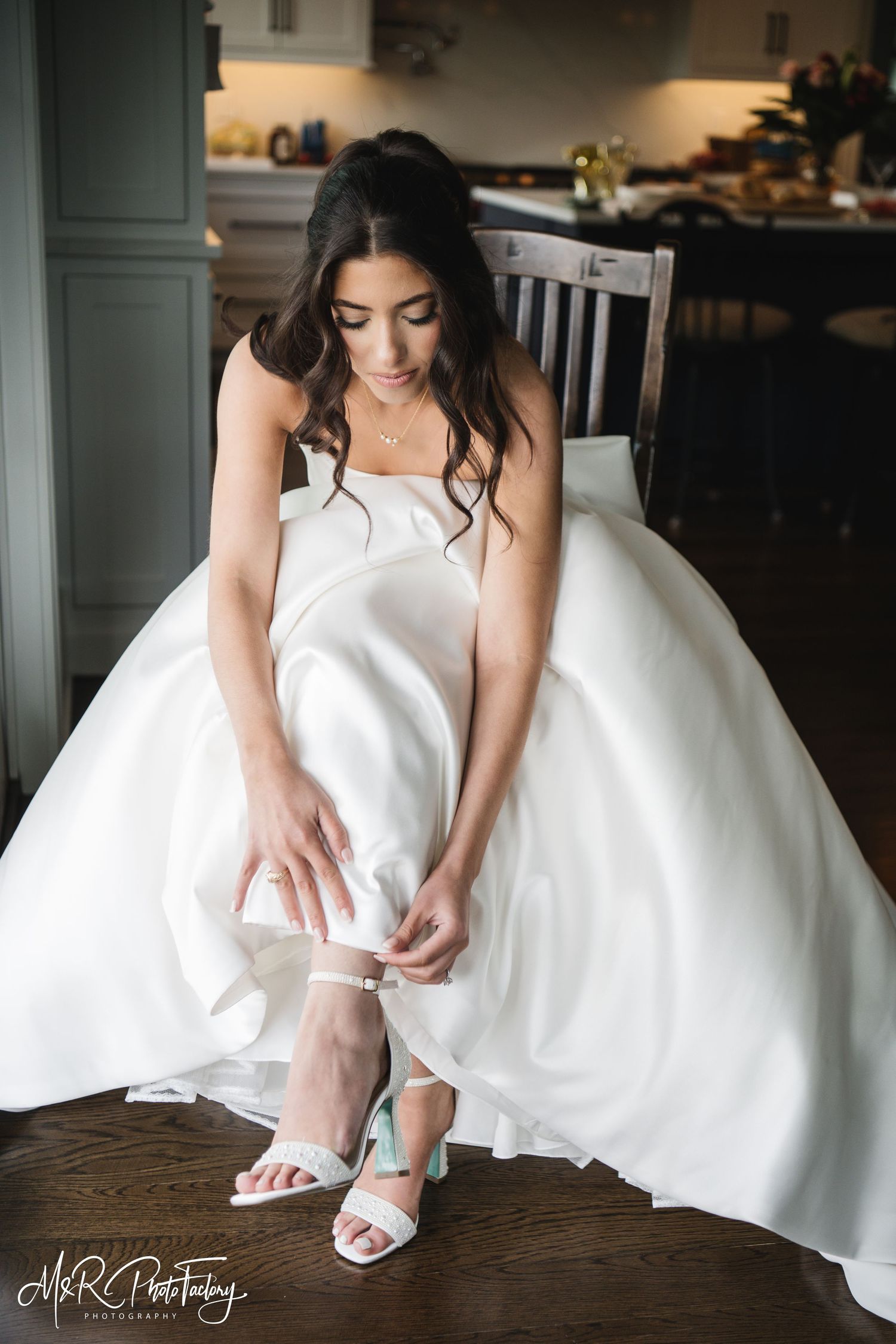 Bride in voluminous white ball gown seated while fastening strappy white bridal heels with blue detail.