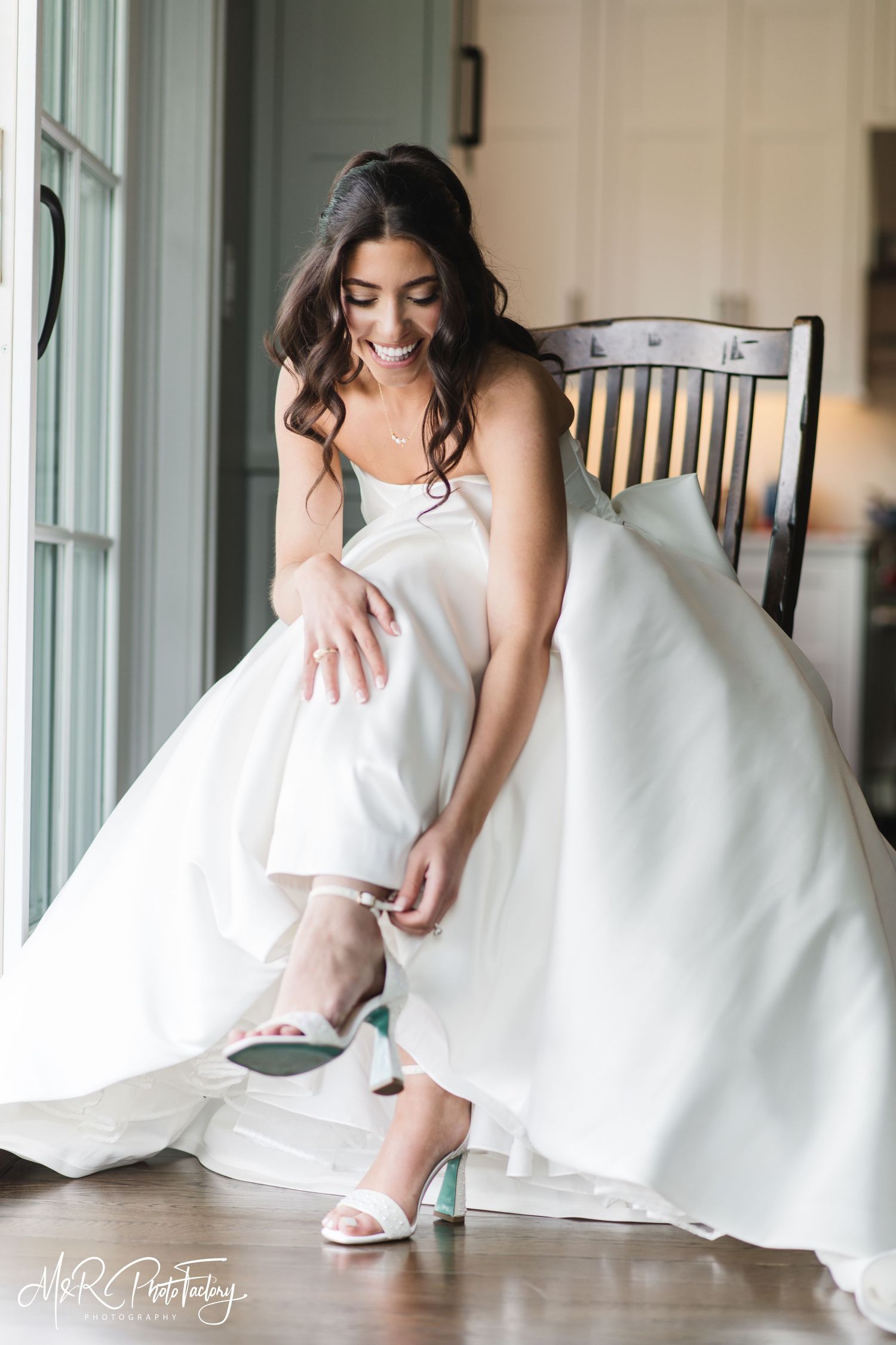 Bride in white wedding gown smiling while putting on elegant heeled wedding shoes.