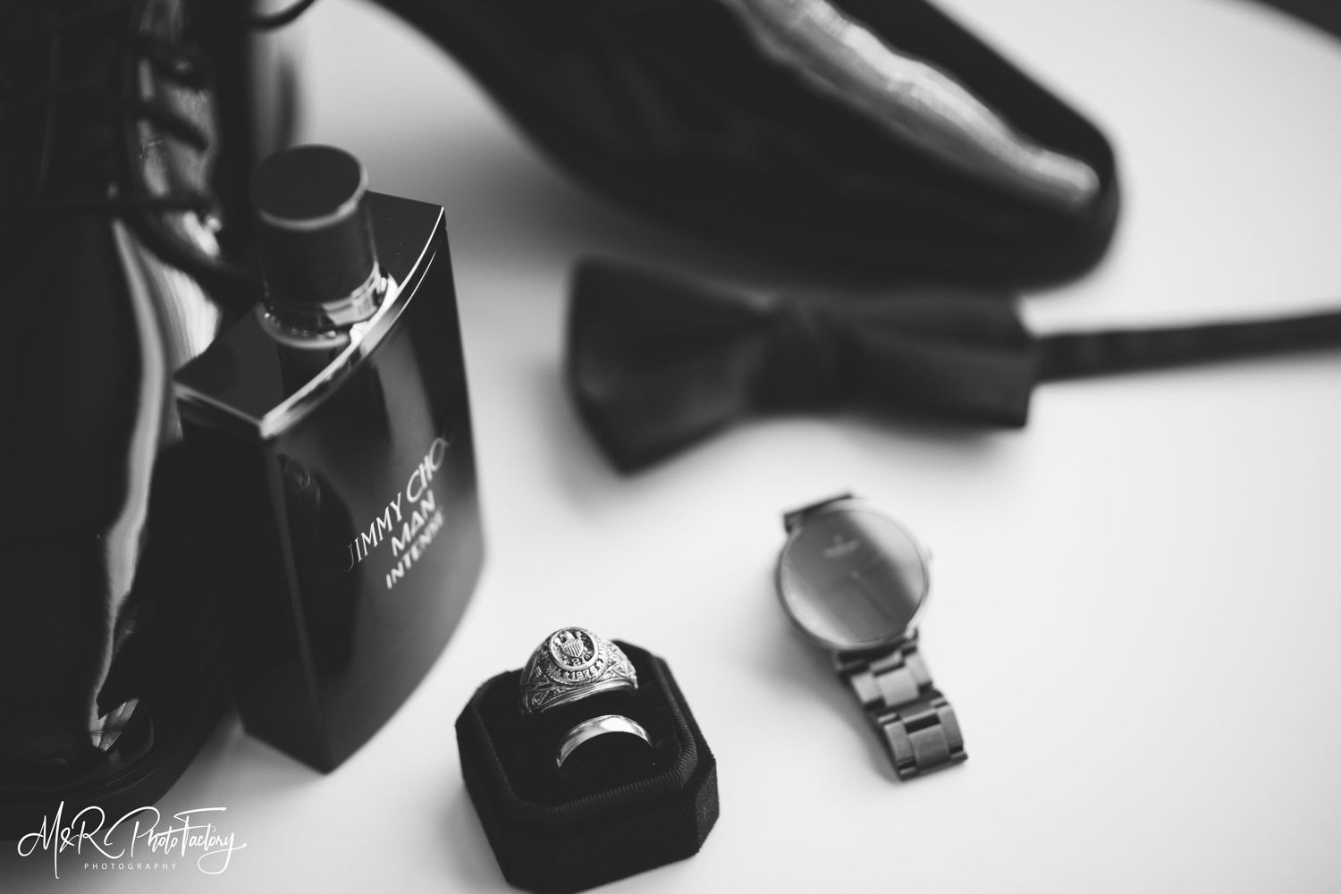 Black and white flat lay of groom accessories including cologne, ring, watch, and bow tie.