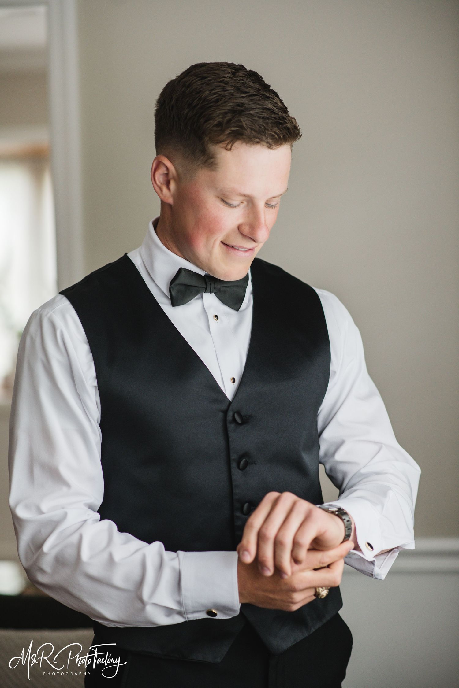 Groom in black vest and bow tie adjusting his cufflinks while smiling.