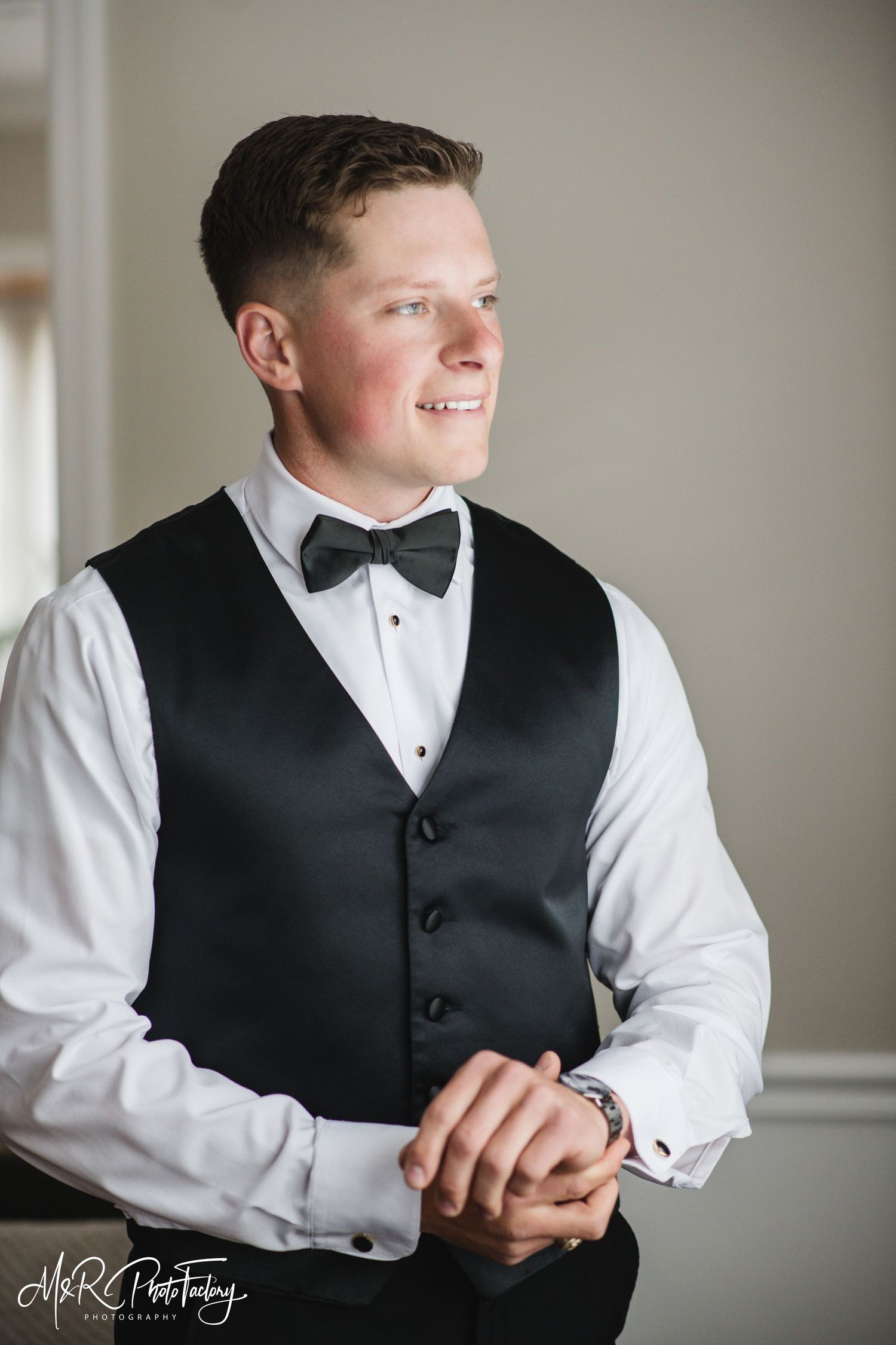 Handsome groom in black vest and bow tie smiling while looking to the side.