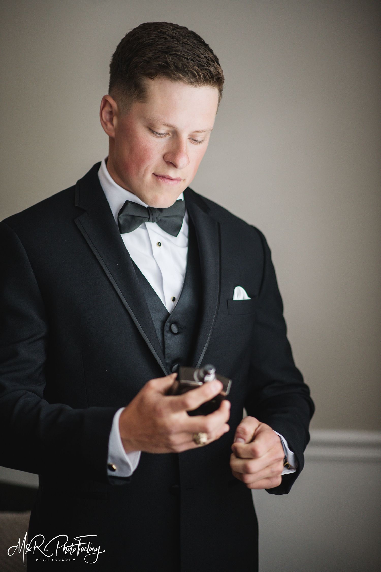 Dapper groom in black tuxedo looking down at cologne bottle before wedding.