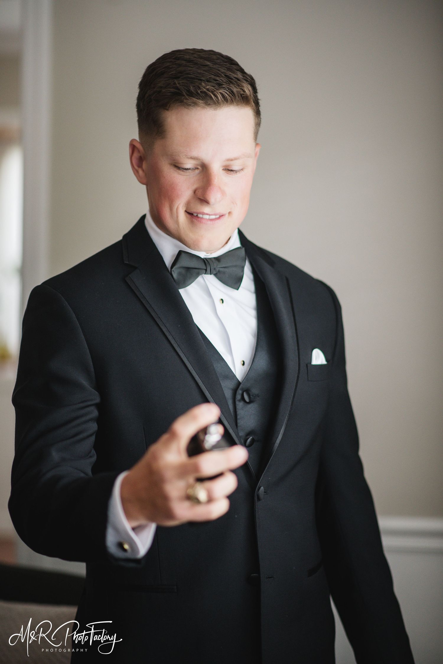 Groom in black tuxedo smiling while holding cologne bottle on wedding day.