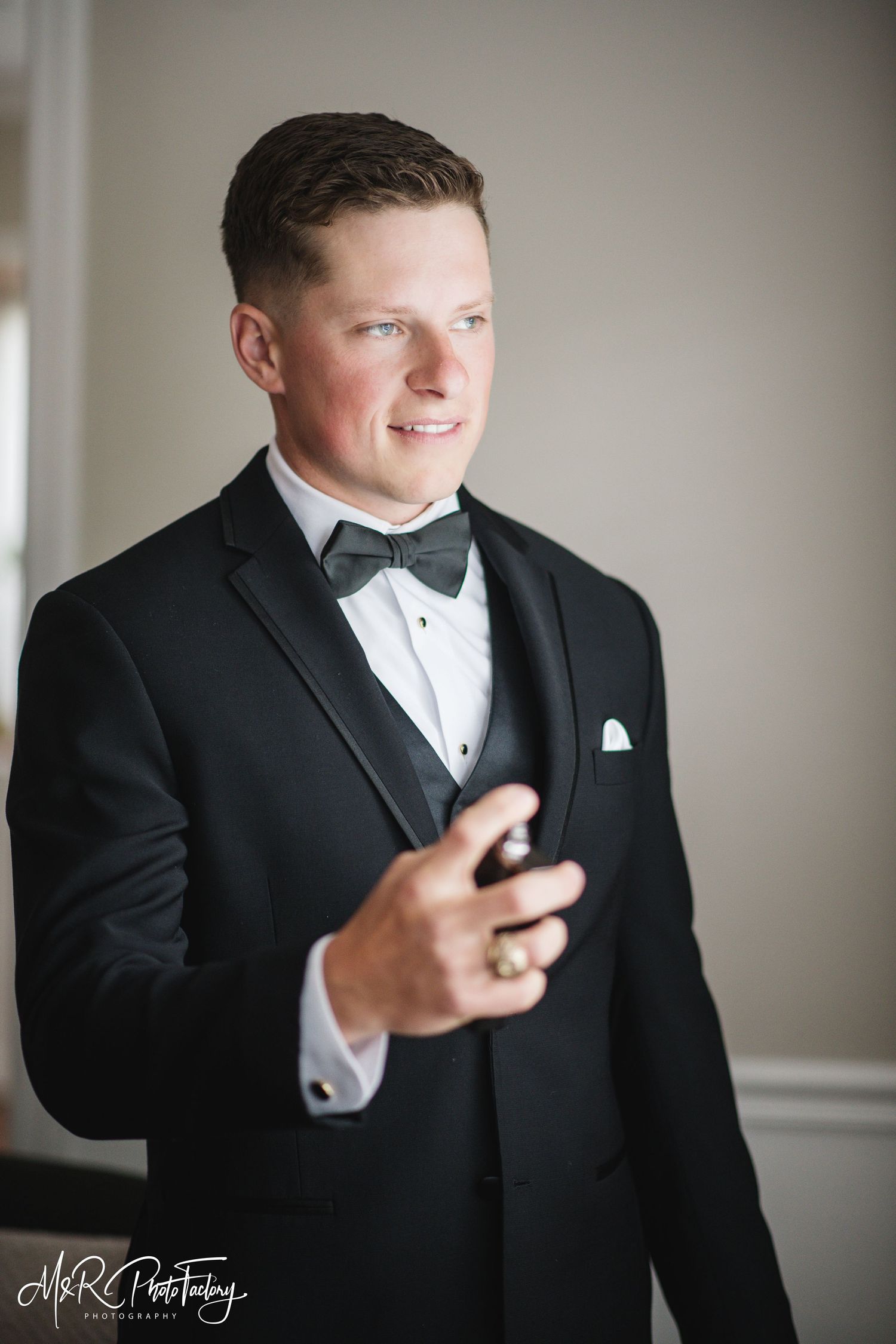 Groom in black tuxedo holding cologne bottle while smiling on wedding day.