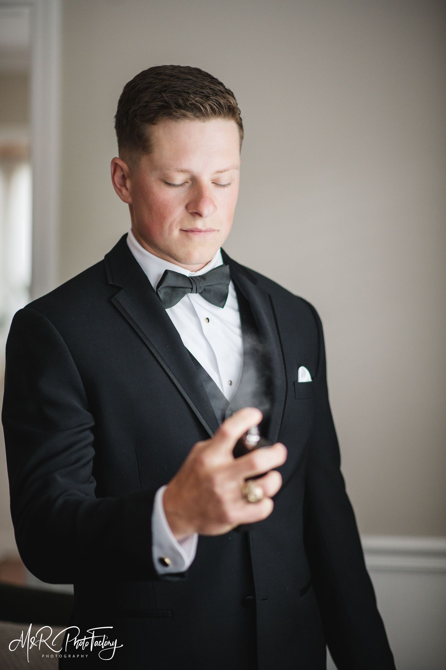 Groom in black tuxedo with bow tie applying cologne before wedding ceremony.