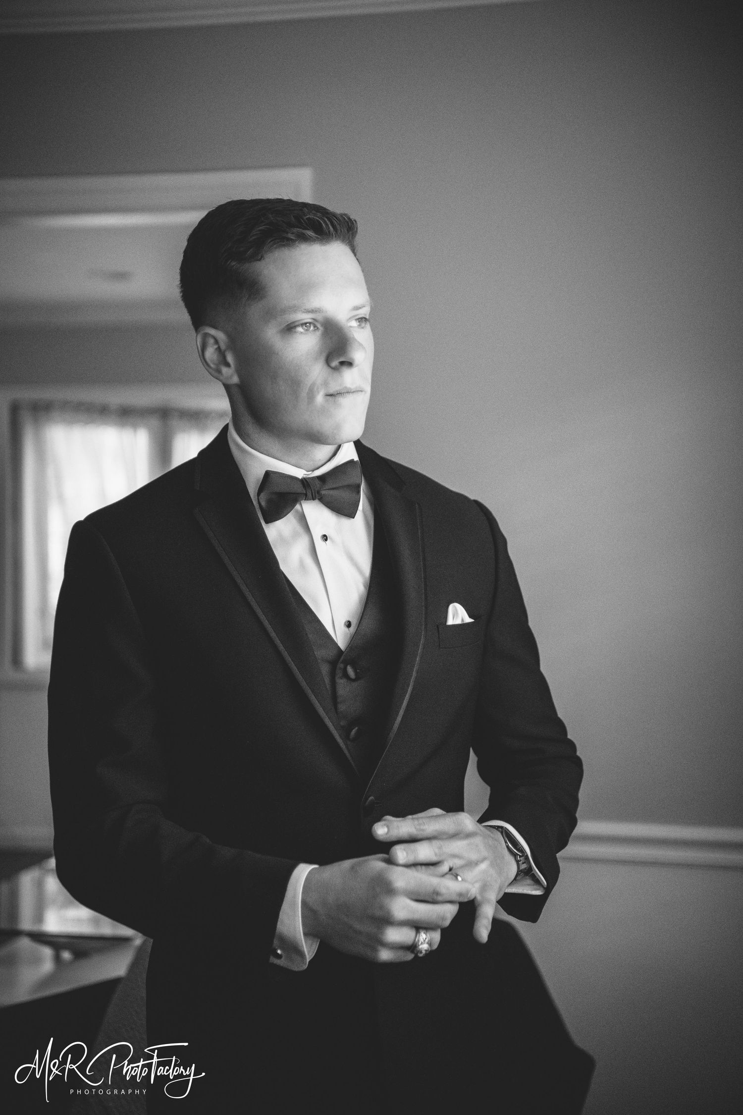 Groom in black tuxedo with bow tie stands thoughtfully in black and white wedding portrait.