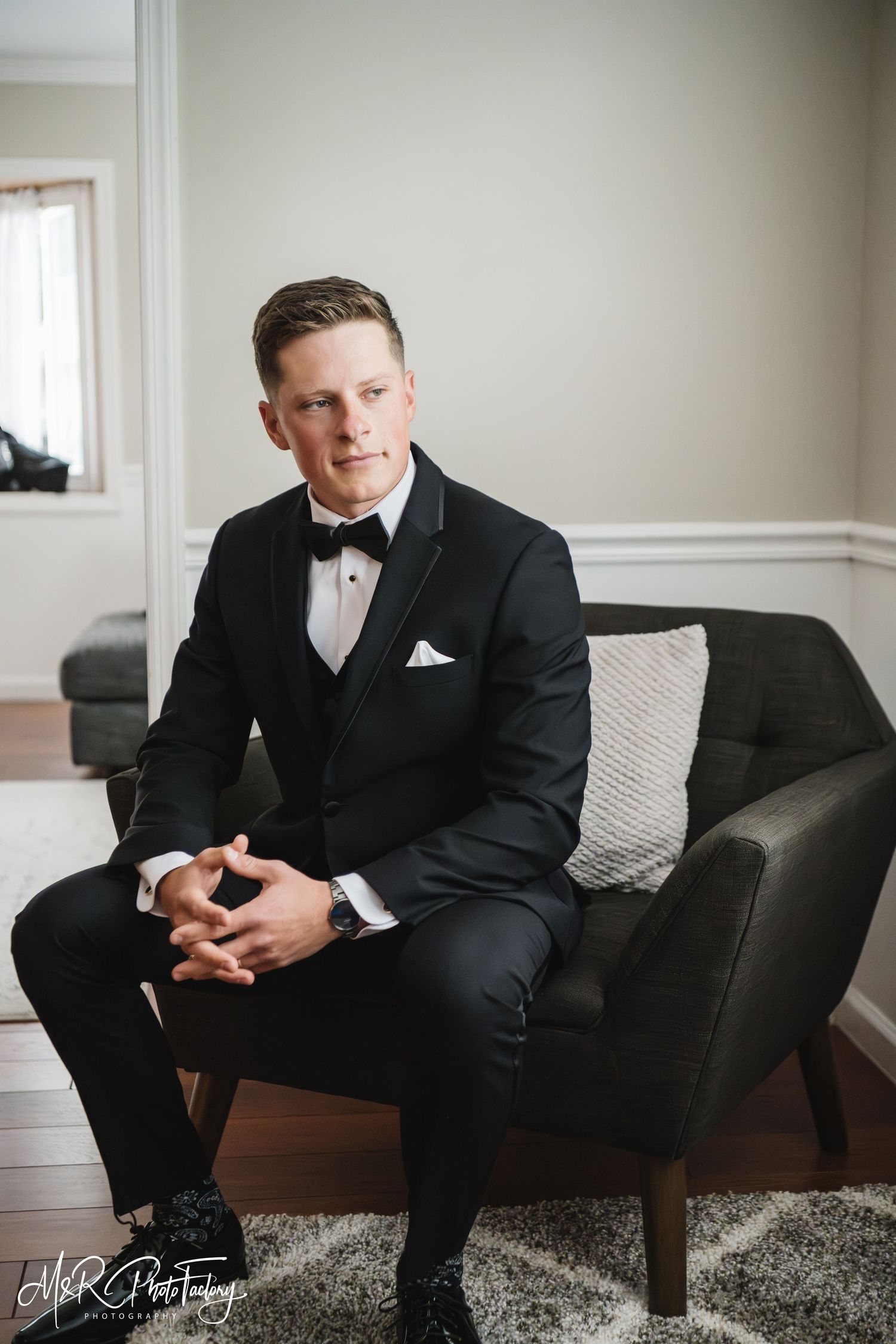 Groom in black tuxedo sits confidently in armchair in a color wedding portrait.