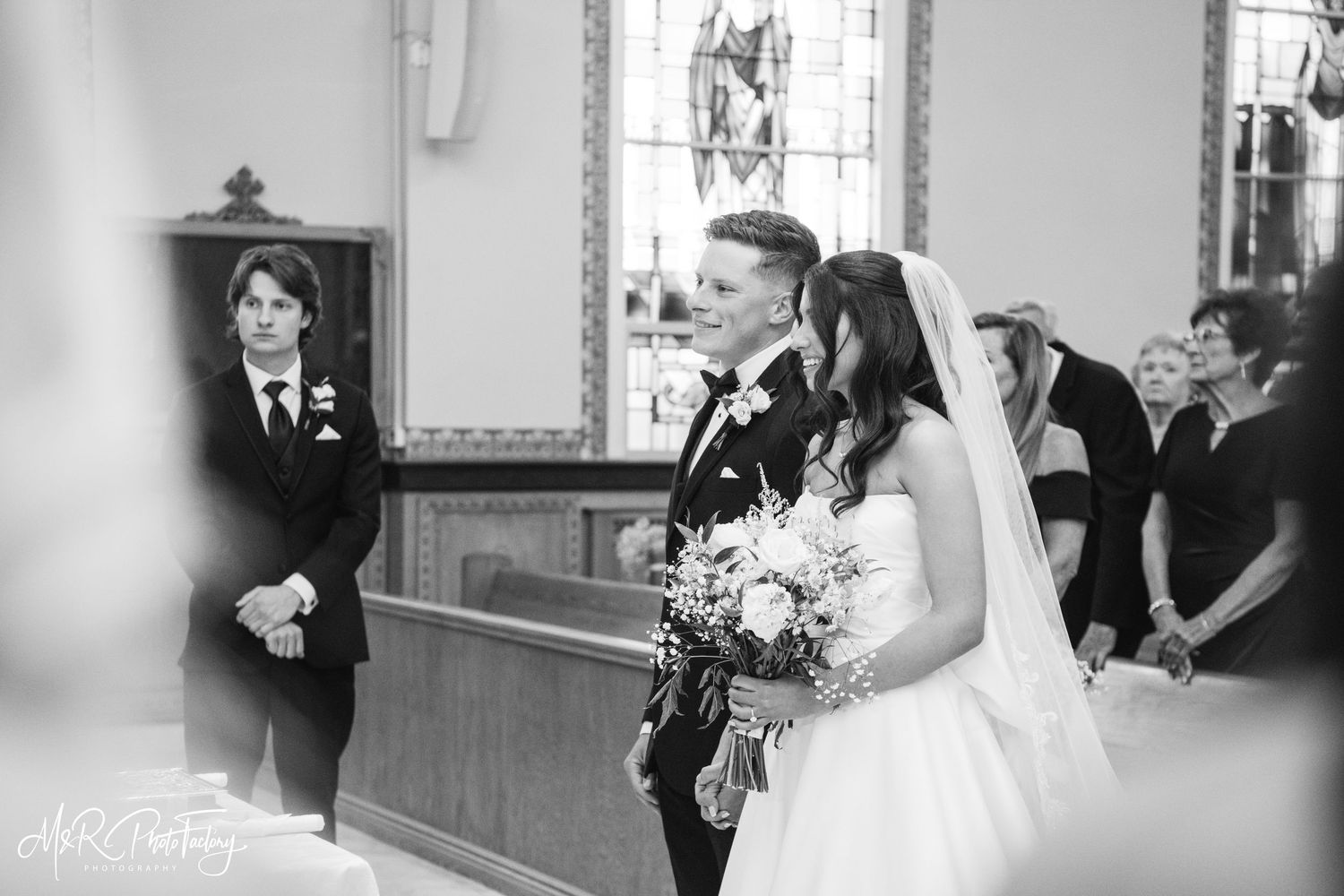 Bride holding bouquet and groom stand together at church altar in black and white.