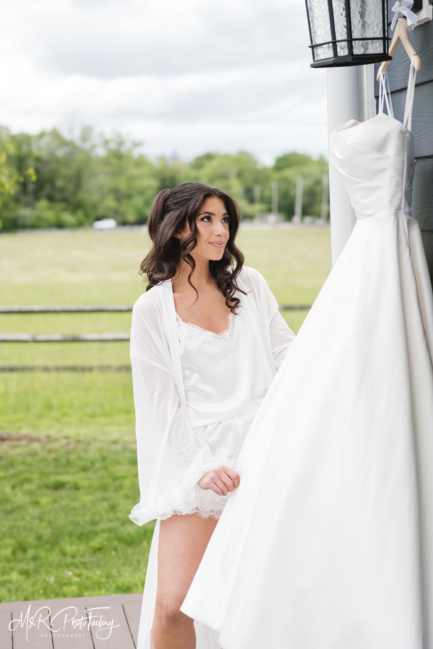 Bride in white robe admiring her strapless wedding dress hanging outdoors at a farm venue.