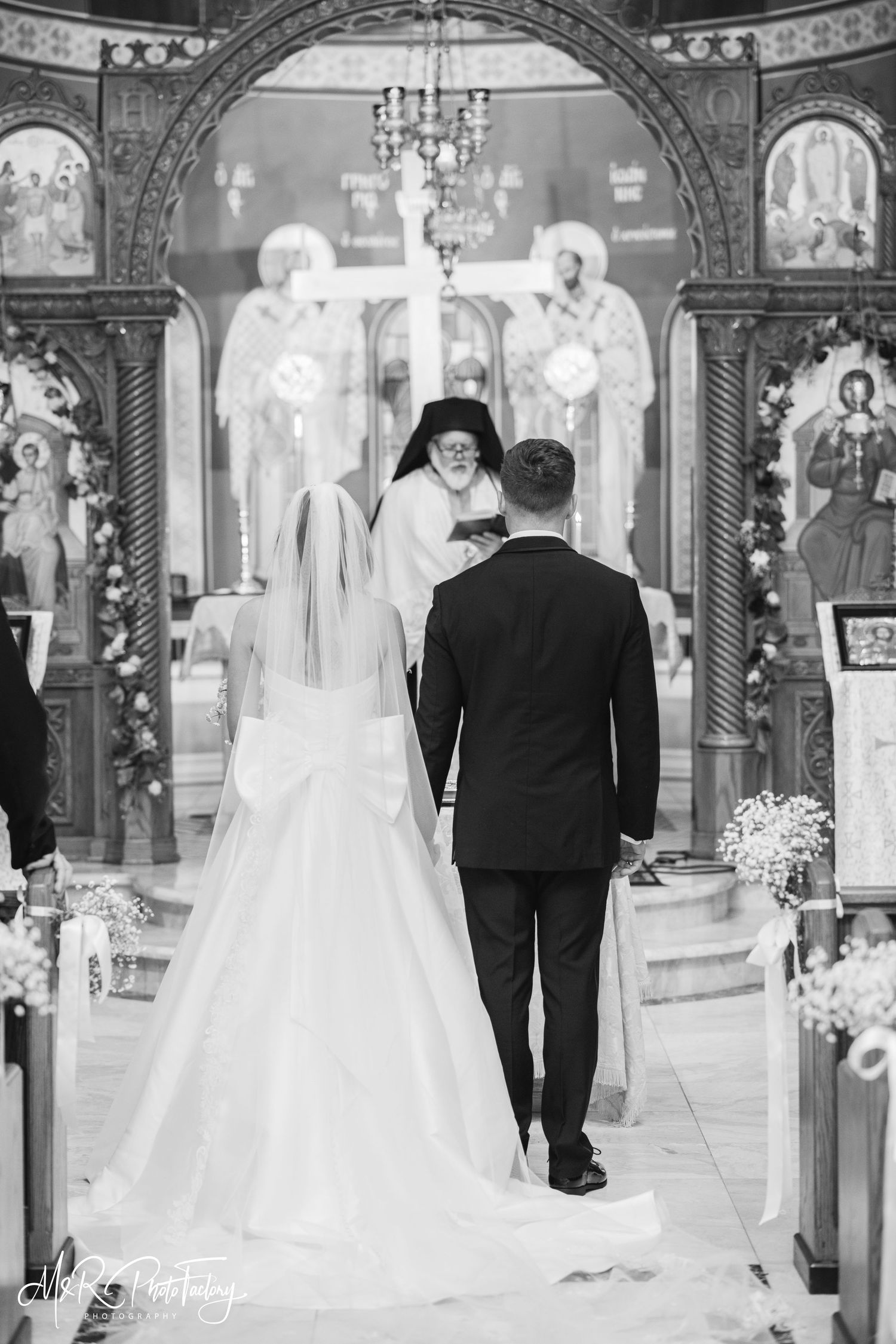 Bride and groom stand before Orthodox priest at ornate church altar in black and white.