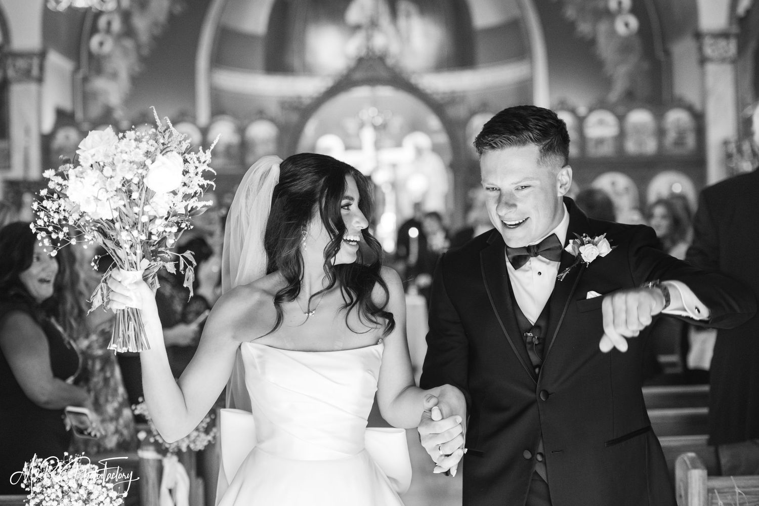 Black and white photo of laughing bride and groom walking down church aisle after wedding ceremony.