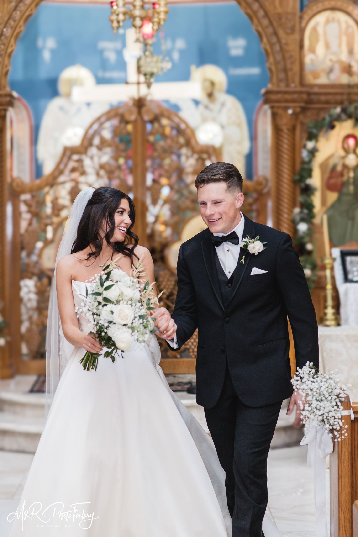 Smiling bride in ball gown and groom in tuxedo walking hand in hand past ornate church altar.