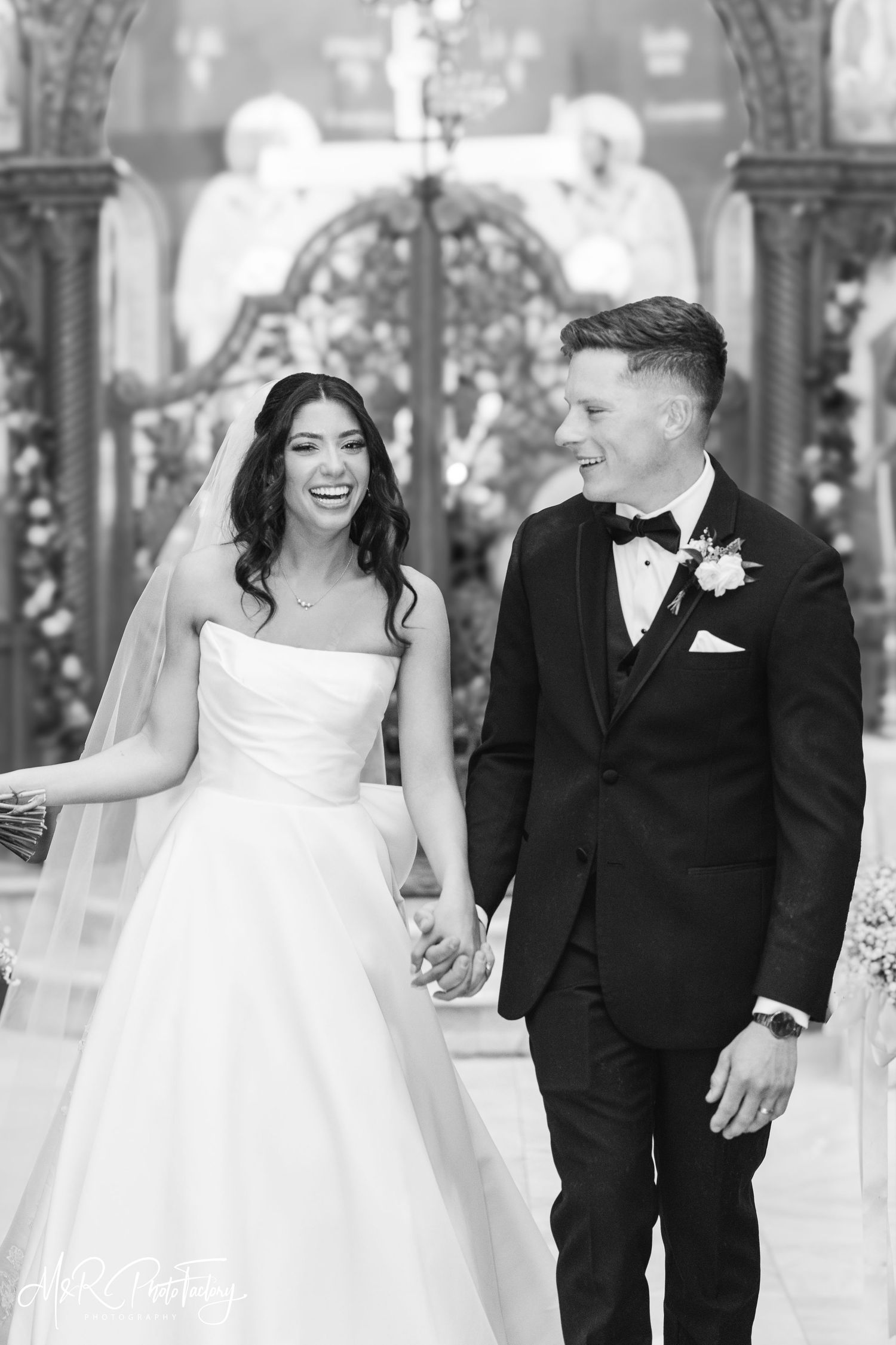 Black and white portrait of radiant bride and groom holding hands and laughing inside decorated church.