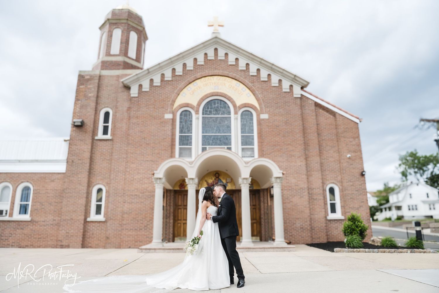 Bride and groom sharing a kiss outside a brick church with arched windows on their wedding day.