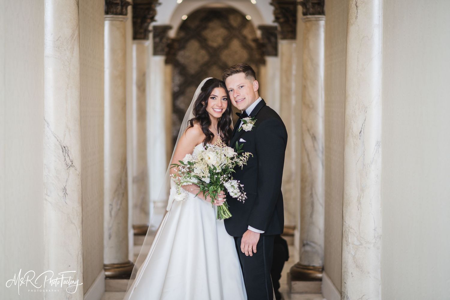 Bride and groom posing together between ornate marble columns holding a white floral wedding bouquet.