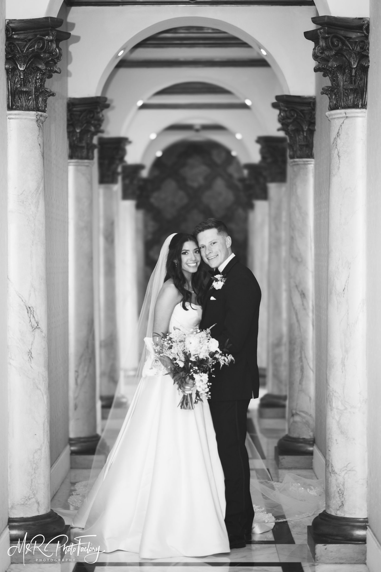 Elegant black and white portrait of bride and groom embracing between marble columns in grand hall.