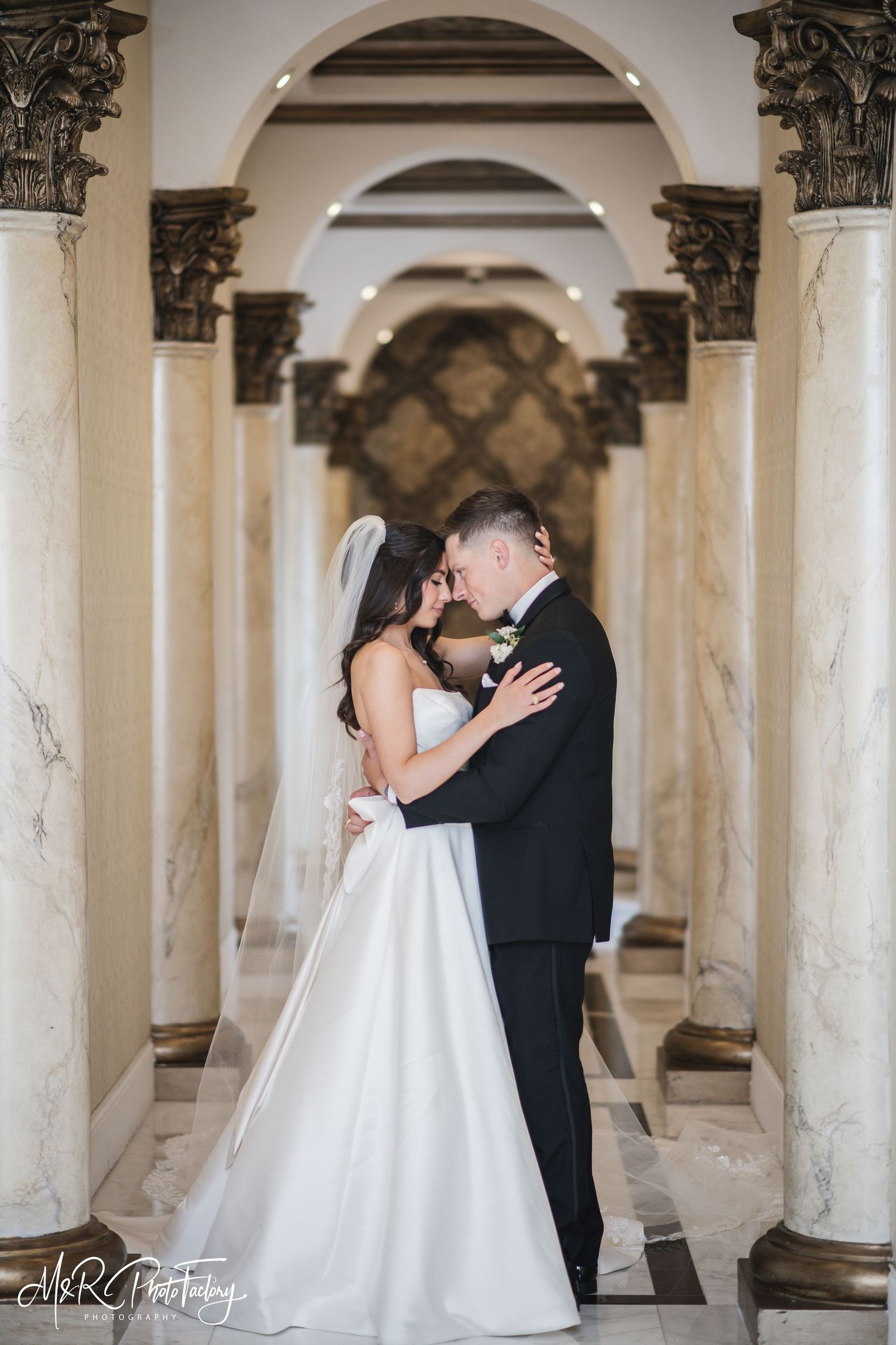 Romantic bride and groom sharing an intimate moment touching foreheads between marble columns in elegant hall.