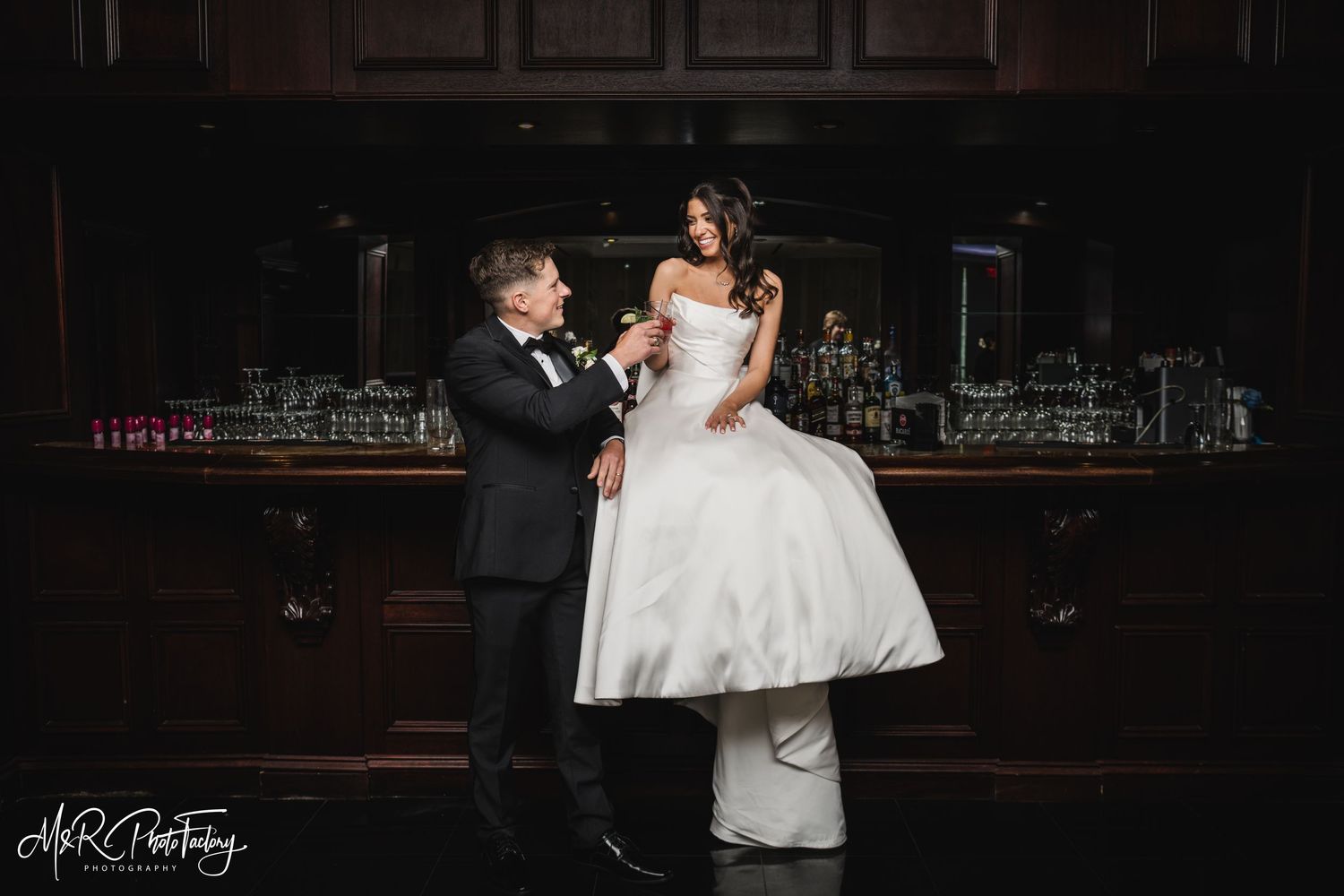 Groom playfully lifting bride in elegant white ball gown at a dimly lit wedding venue bar.