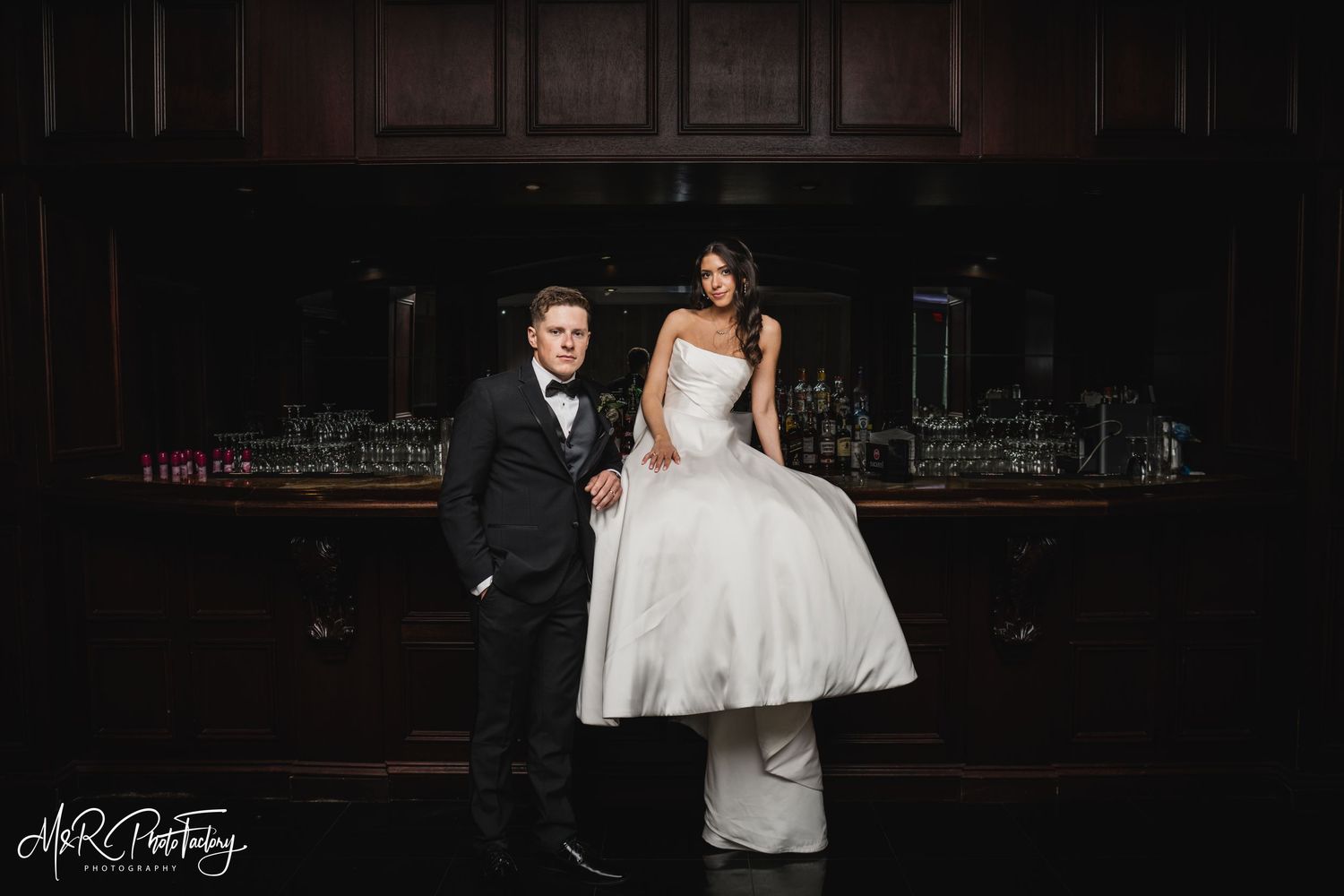 Bride and groom pose elegantly in front of a dark wood bar on their wedding day.