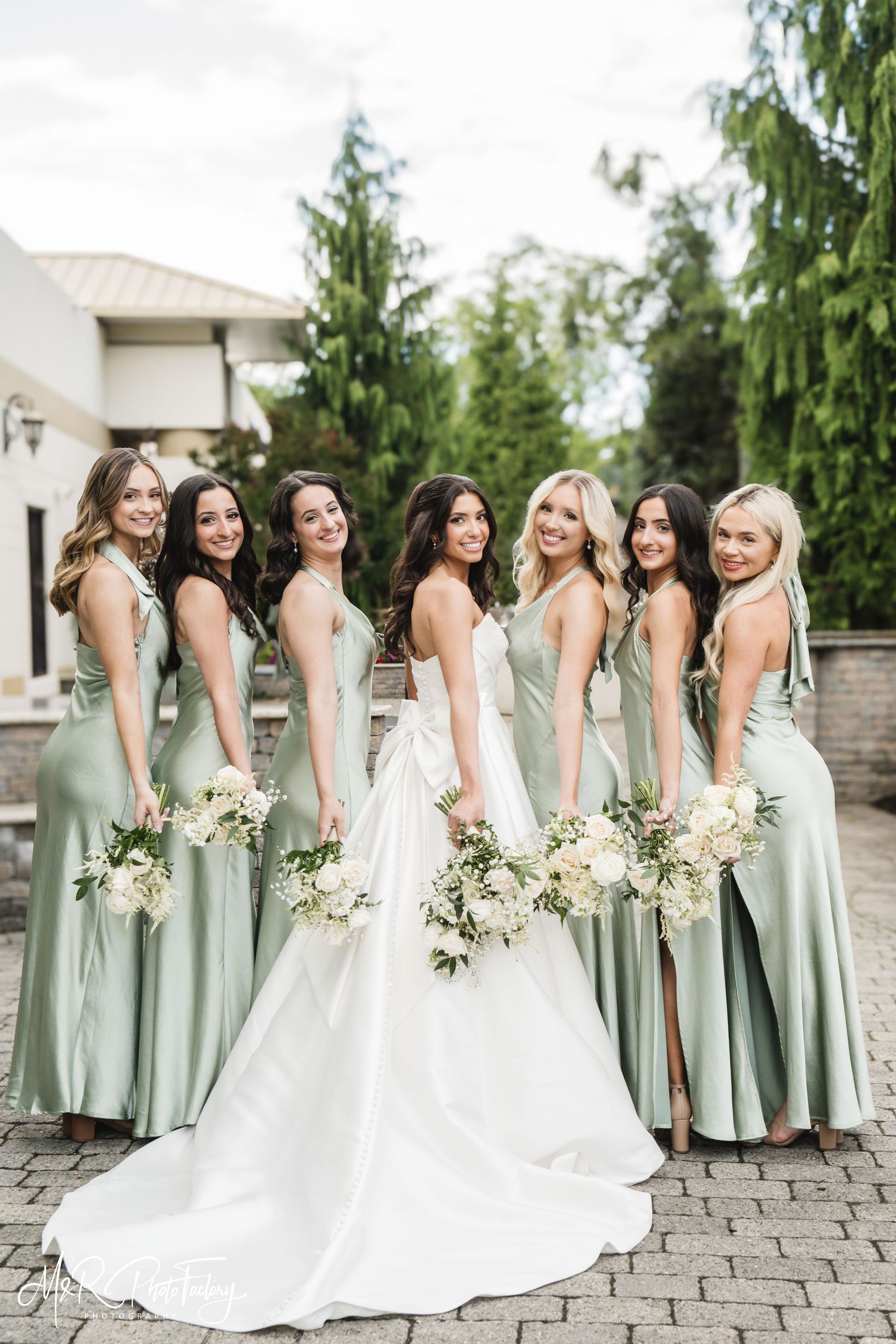 Bride in white gown stands with six bridesmaids in sage green satin dresses holding white bouquets.