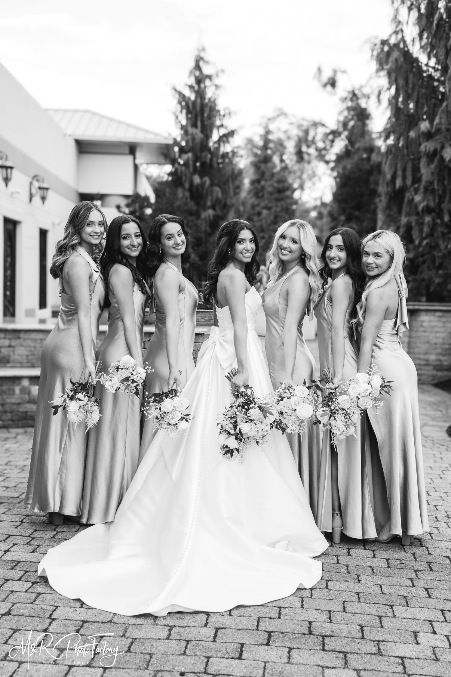 Black and white photo of bride and six bridesmaids in satin gowns holding floral bouquets outdoors.