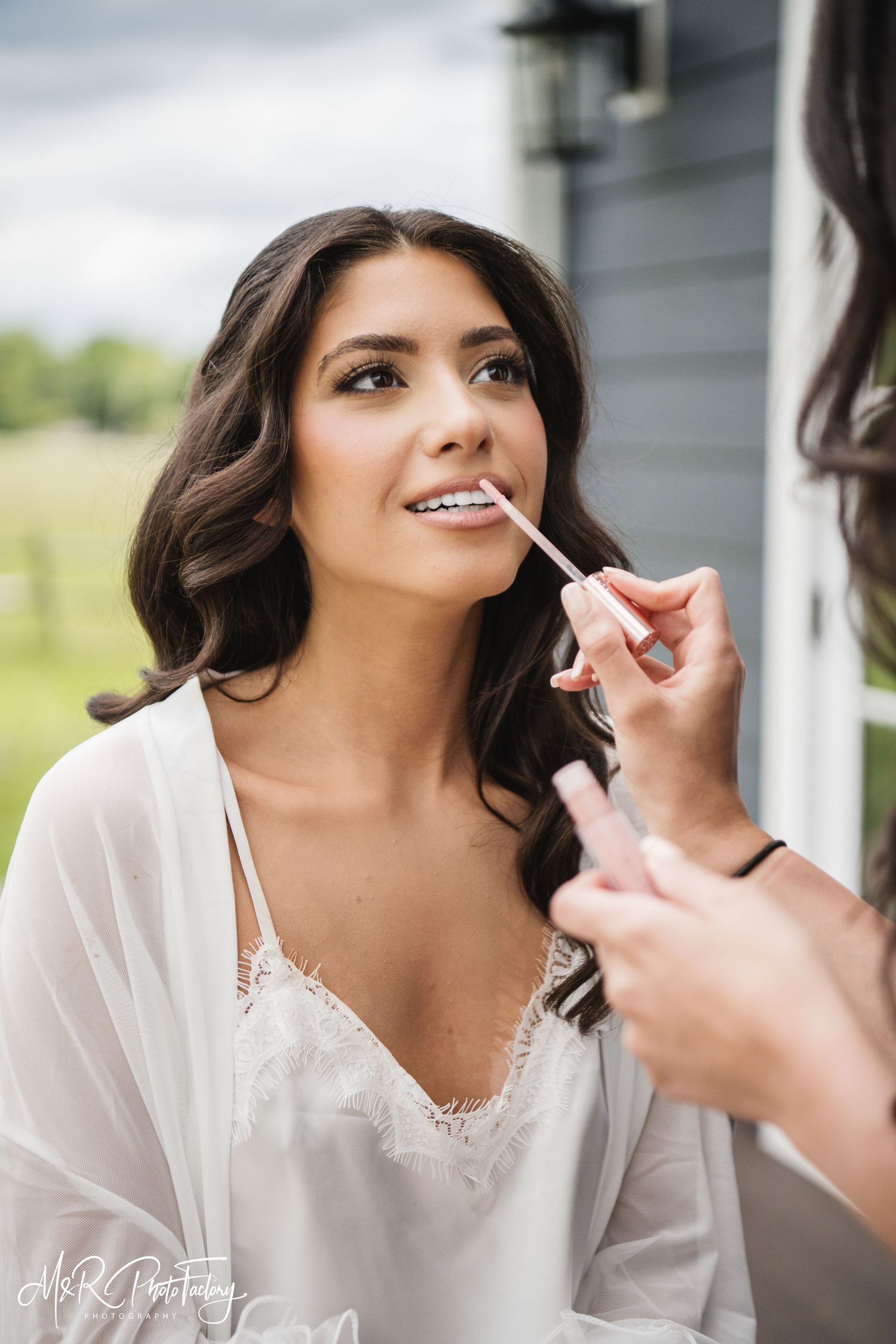 Makeup artist carefully applying lipstick to a beautiful dark-haired bride in a white robe.
