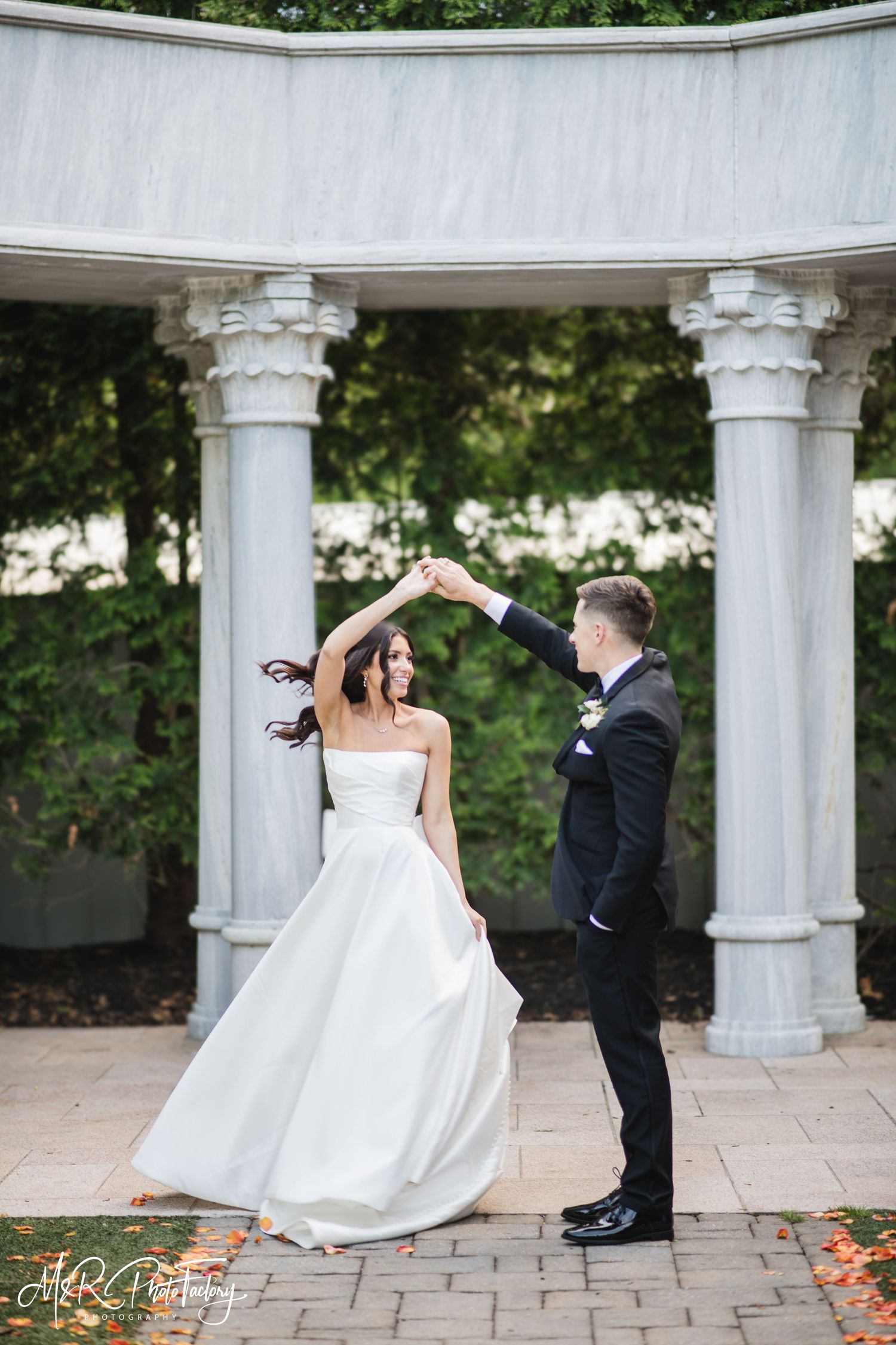 Groom spins bride in a flowing white gown beneath a beautiful stone colonnade on their wedding day.