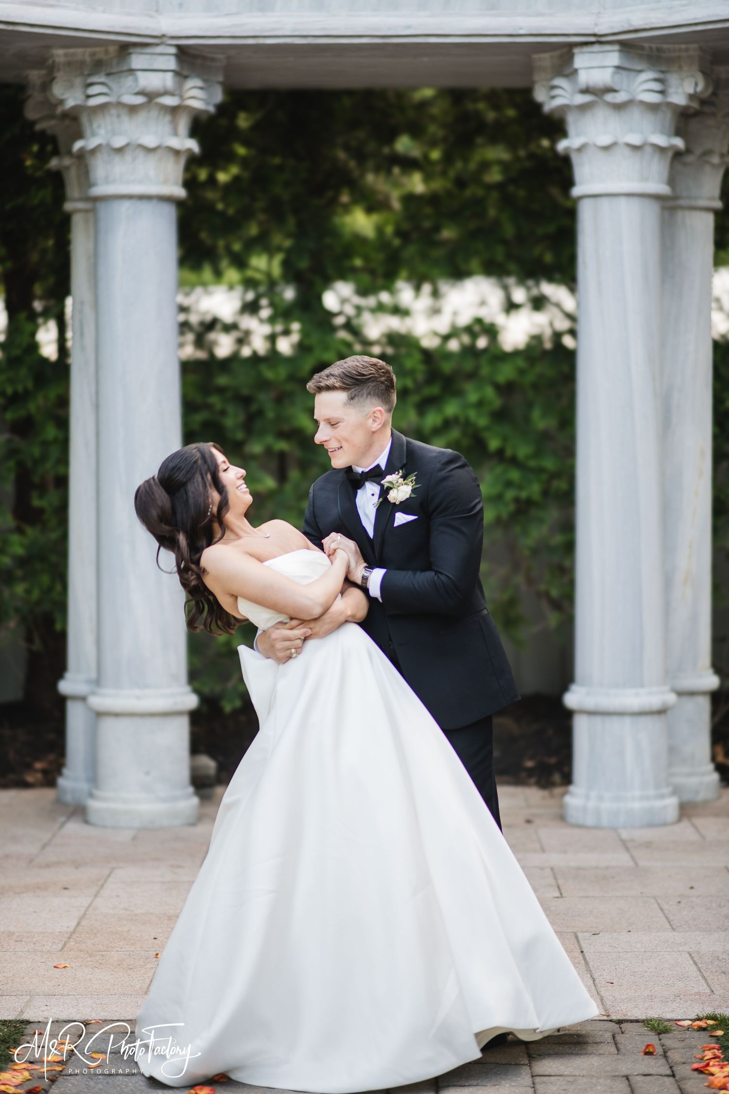 Groom dips bride in a classic white ball gown beside marble columns during romantic wedding portraits.