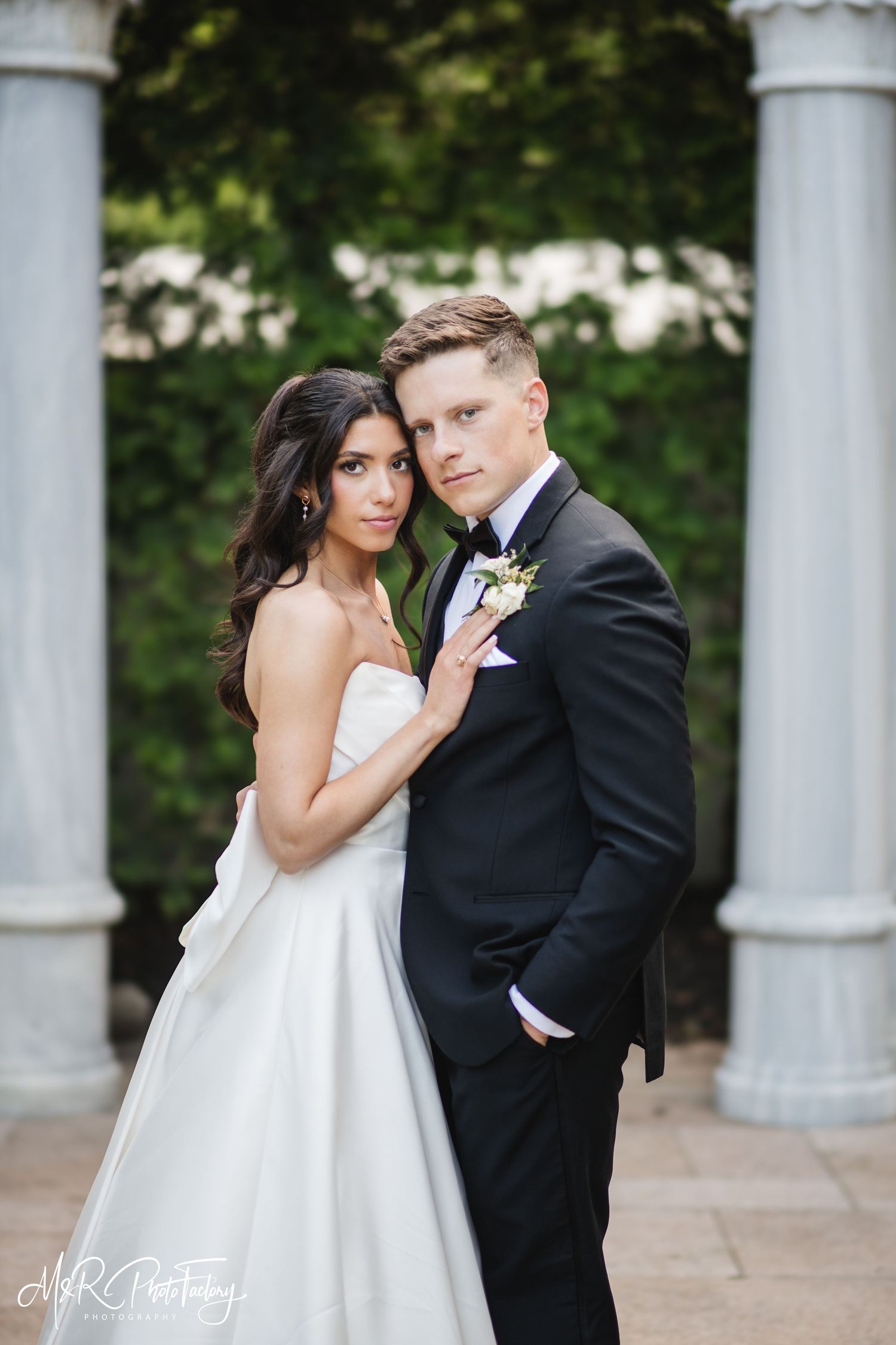 Bride in strapless white gown and groom in black tuxedo pose elegantly before marble columns and lush greenery.