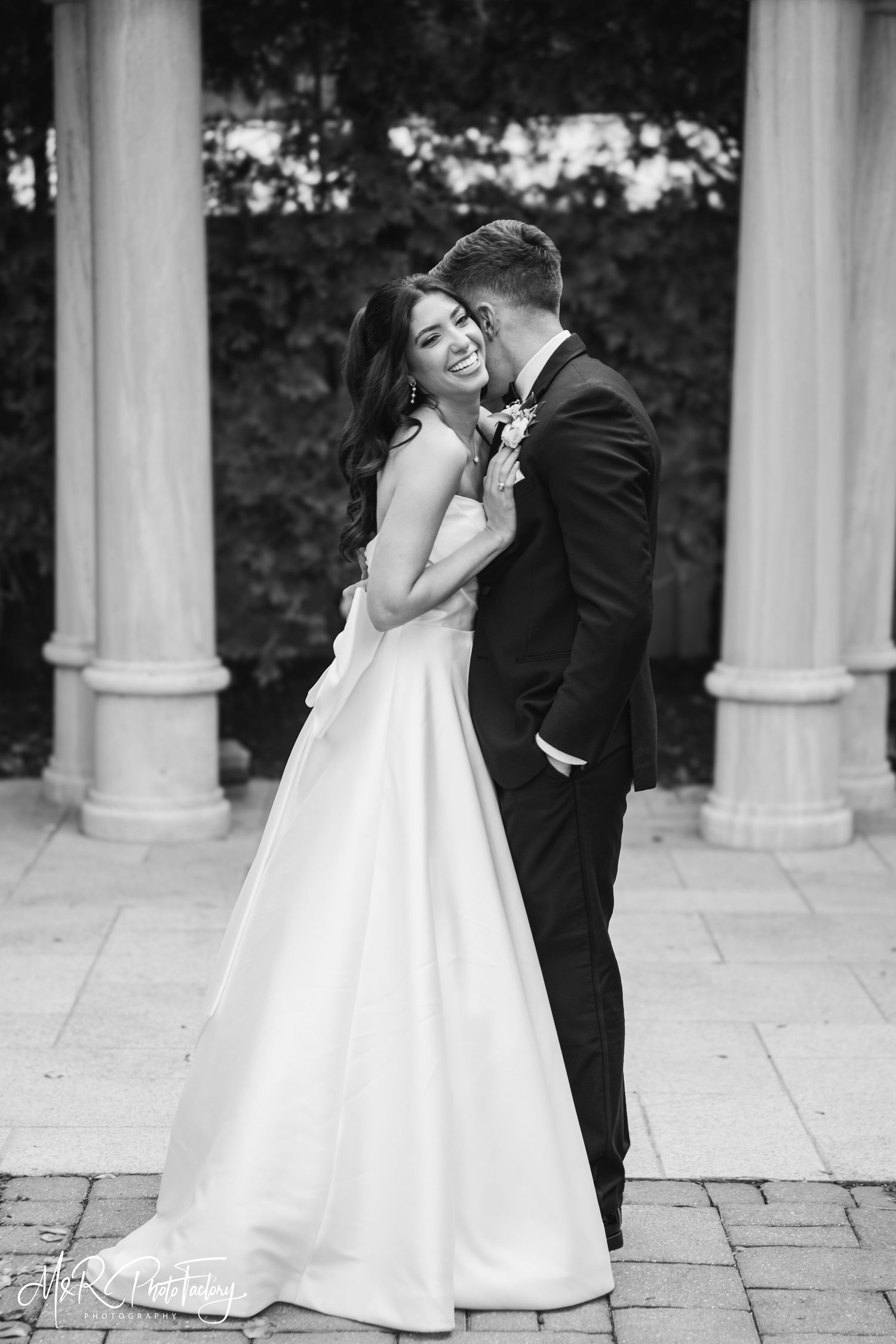Black and white portrait of laughing bride embraced by groom beside elegant stone columns outdoors.