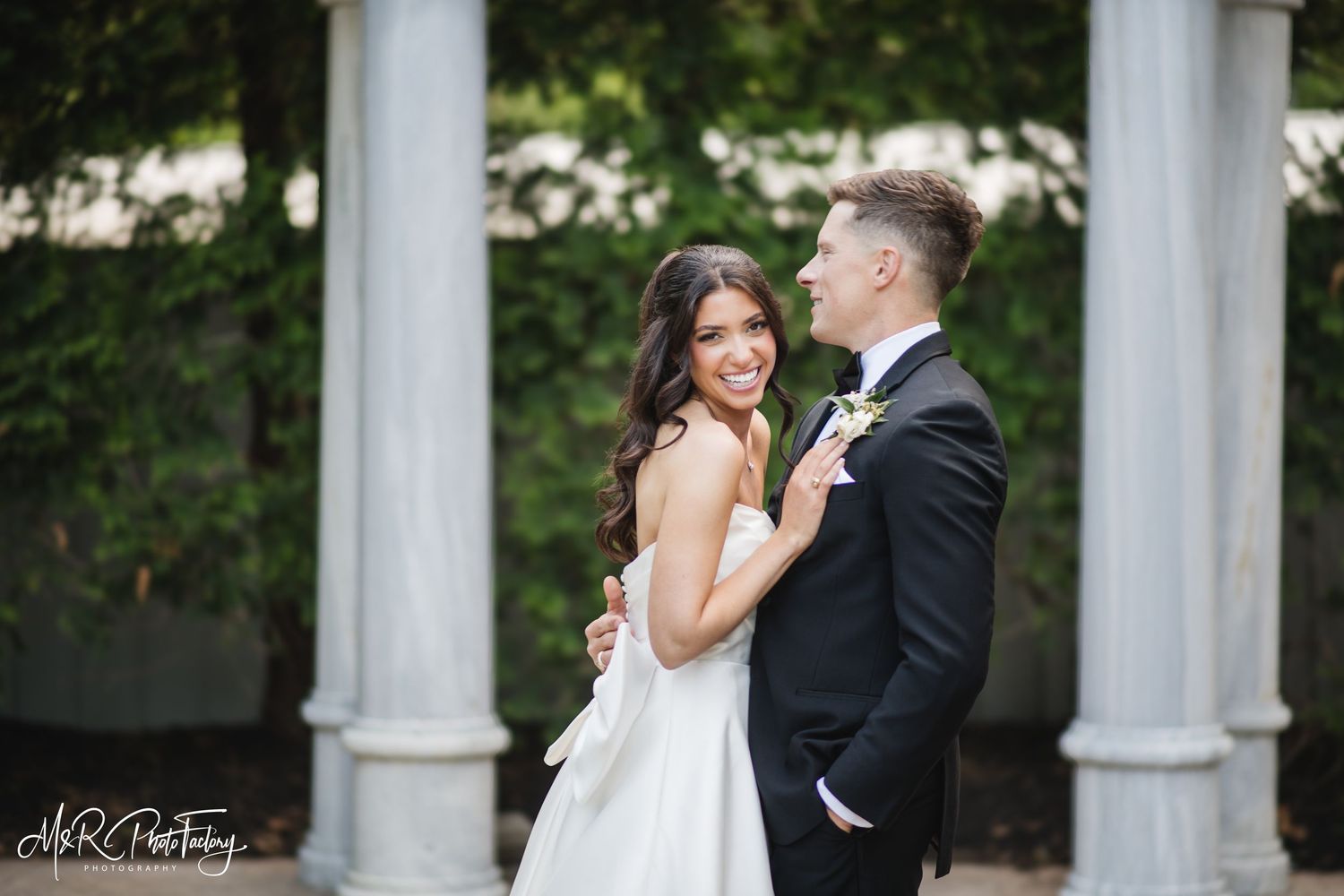Smiling bride in white strapless gown embraces groom in black tuxedo near classic marble columns outdoors.