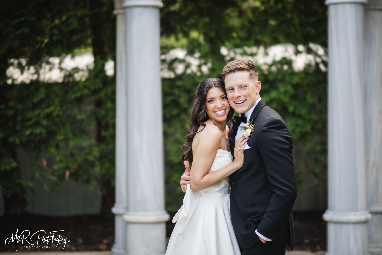 Happy bride and groom share a joyful moment together near elegant marble columns surrounded by green foliage.