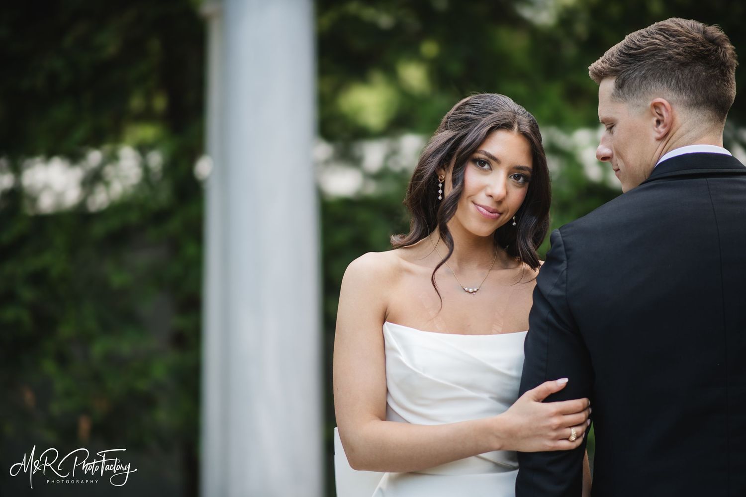 Beautiful dark-haired bride in white strapless gown gazes back while holding grooms arm at outdoor venue.