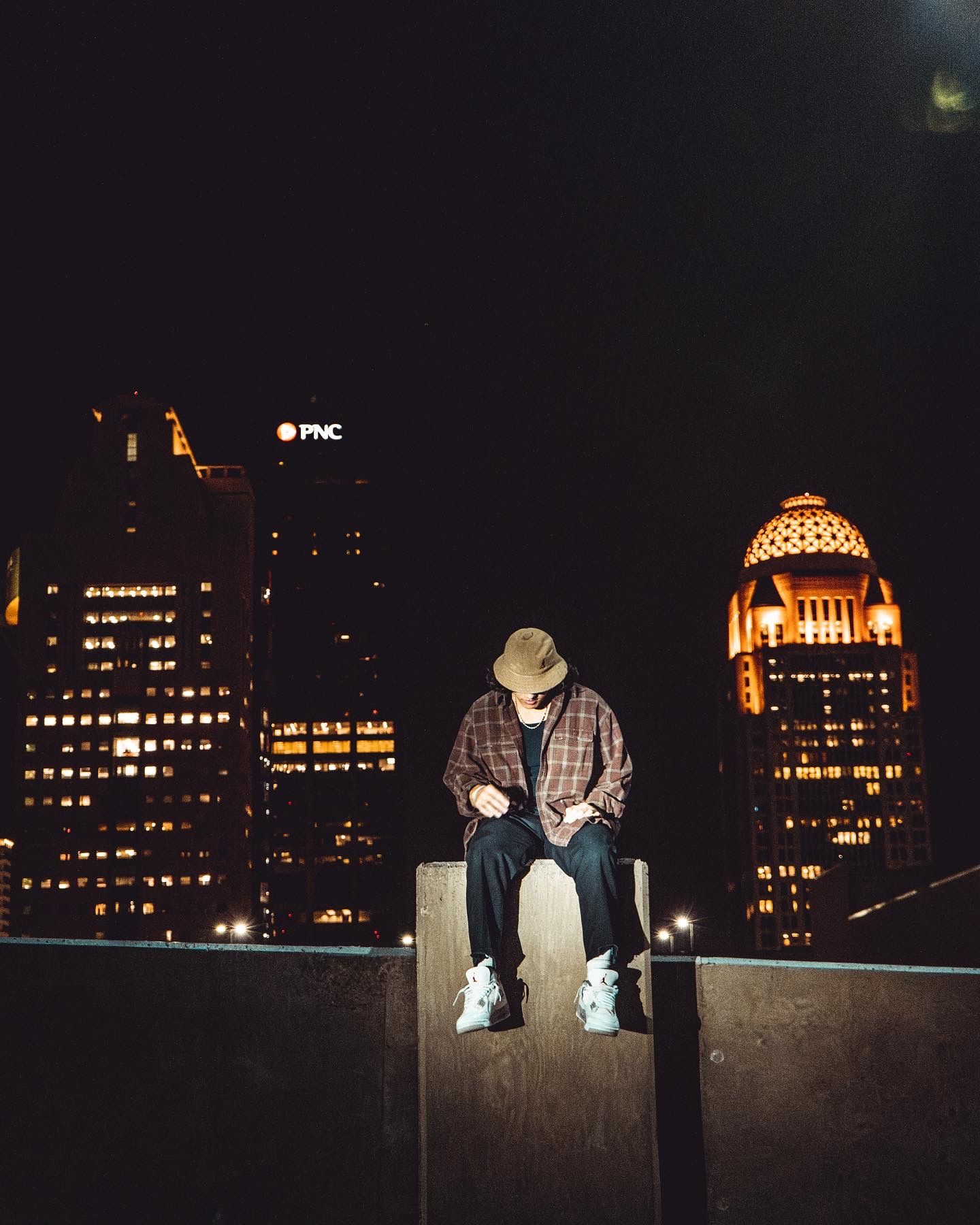 Urban nightscape with illuminated buildings and figure observing from rooftop edge.