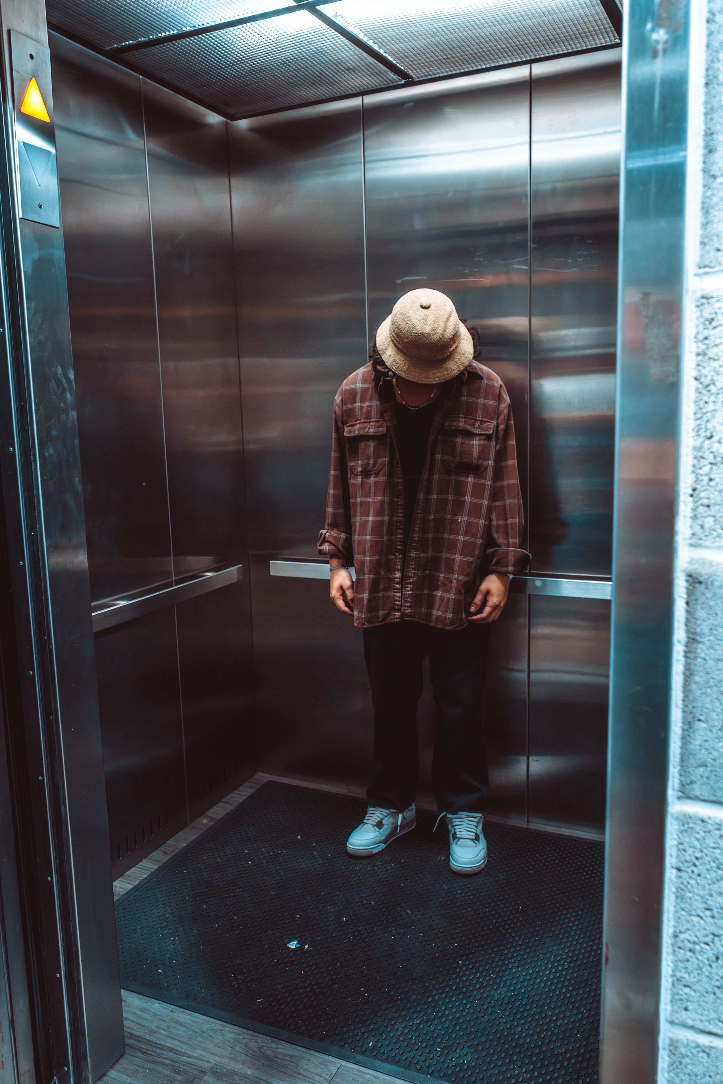 A dark elevator interior with someone wearing a brown plaid shirt and blue shoes standing in the corner.