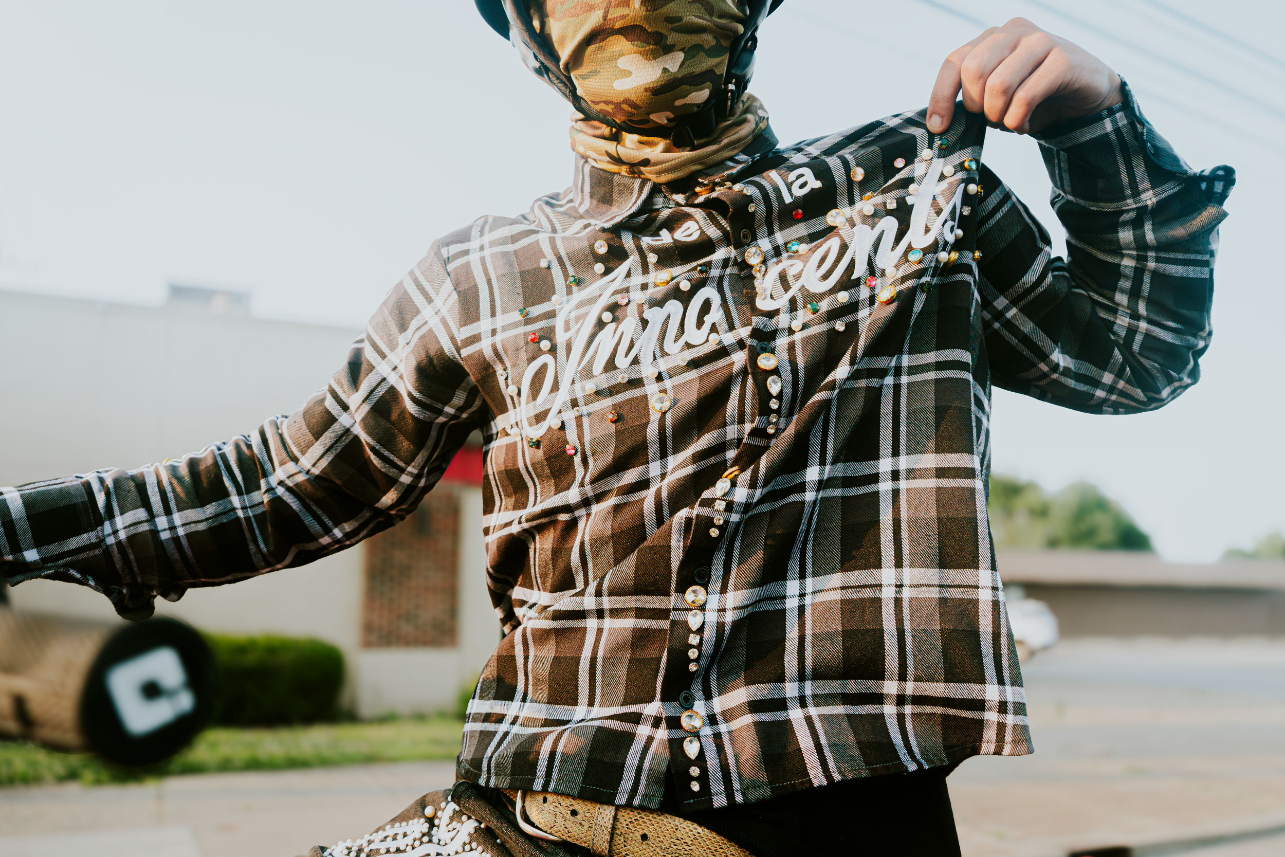 Person wearing plaid flannel shirt and jeans sits casually while playing acoustic guitar outdoors.
