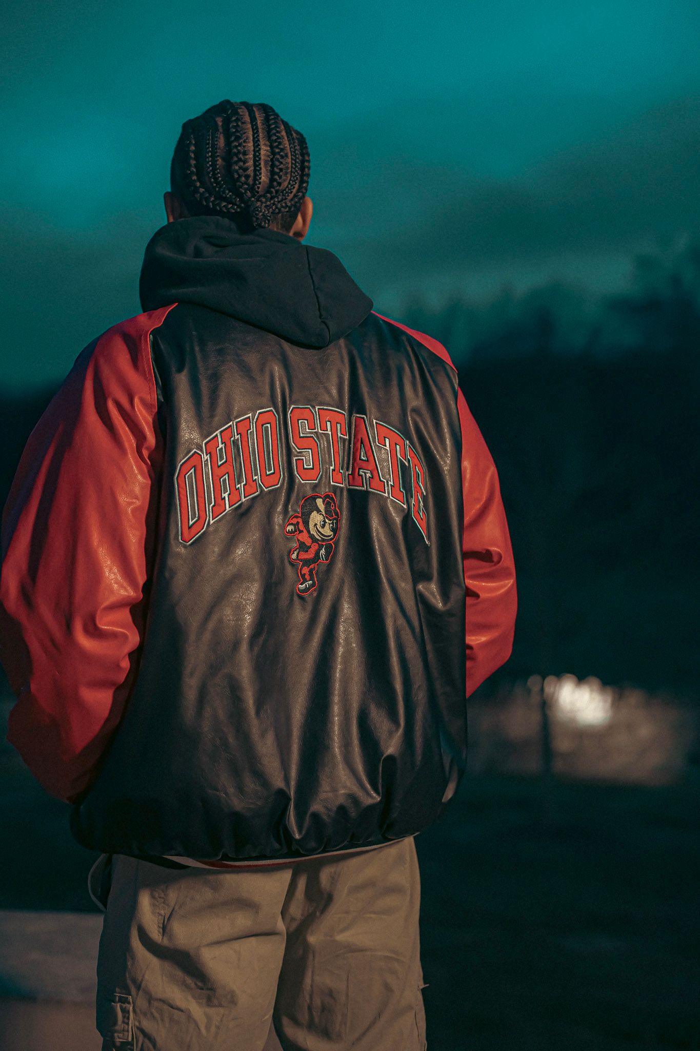 Person wearing an Ohio State varsity jacket with red sleeves stands against a dark turquoise evening sky.