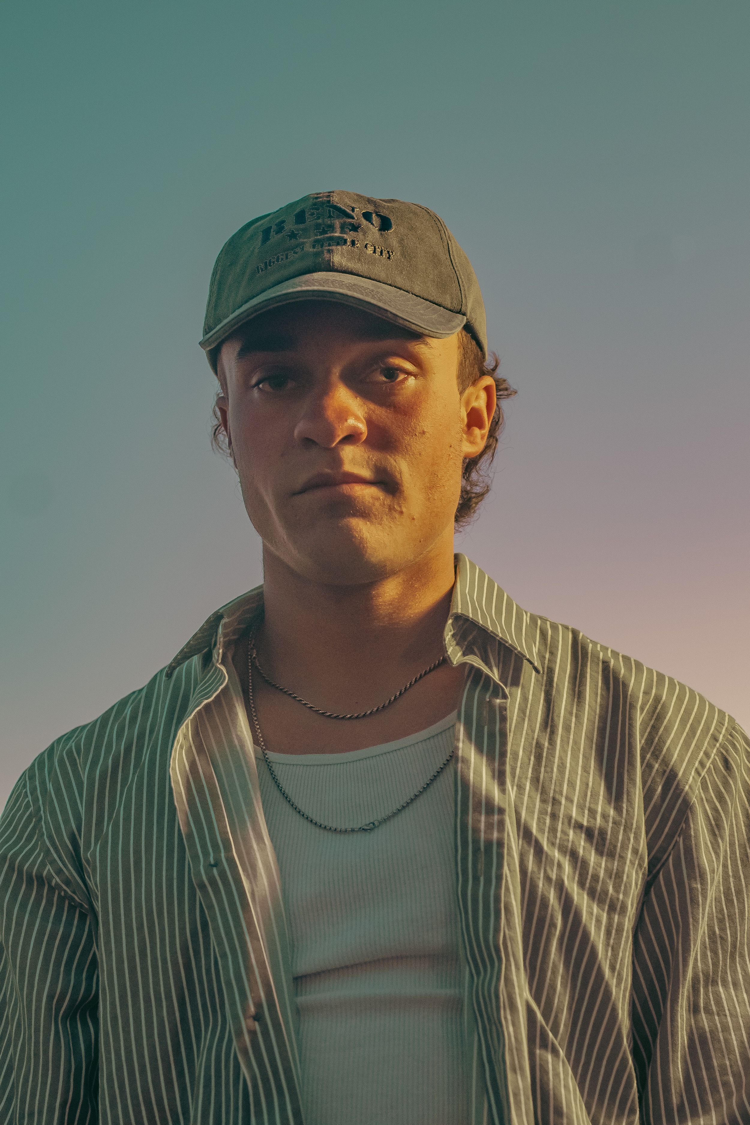 Portrait photograph of someone wearing a baseball cap and striped button-down shirt during golden hour lighting.