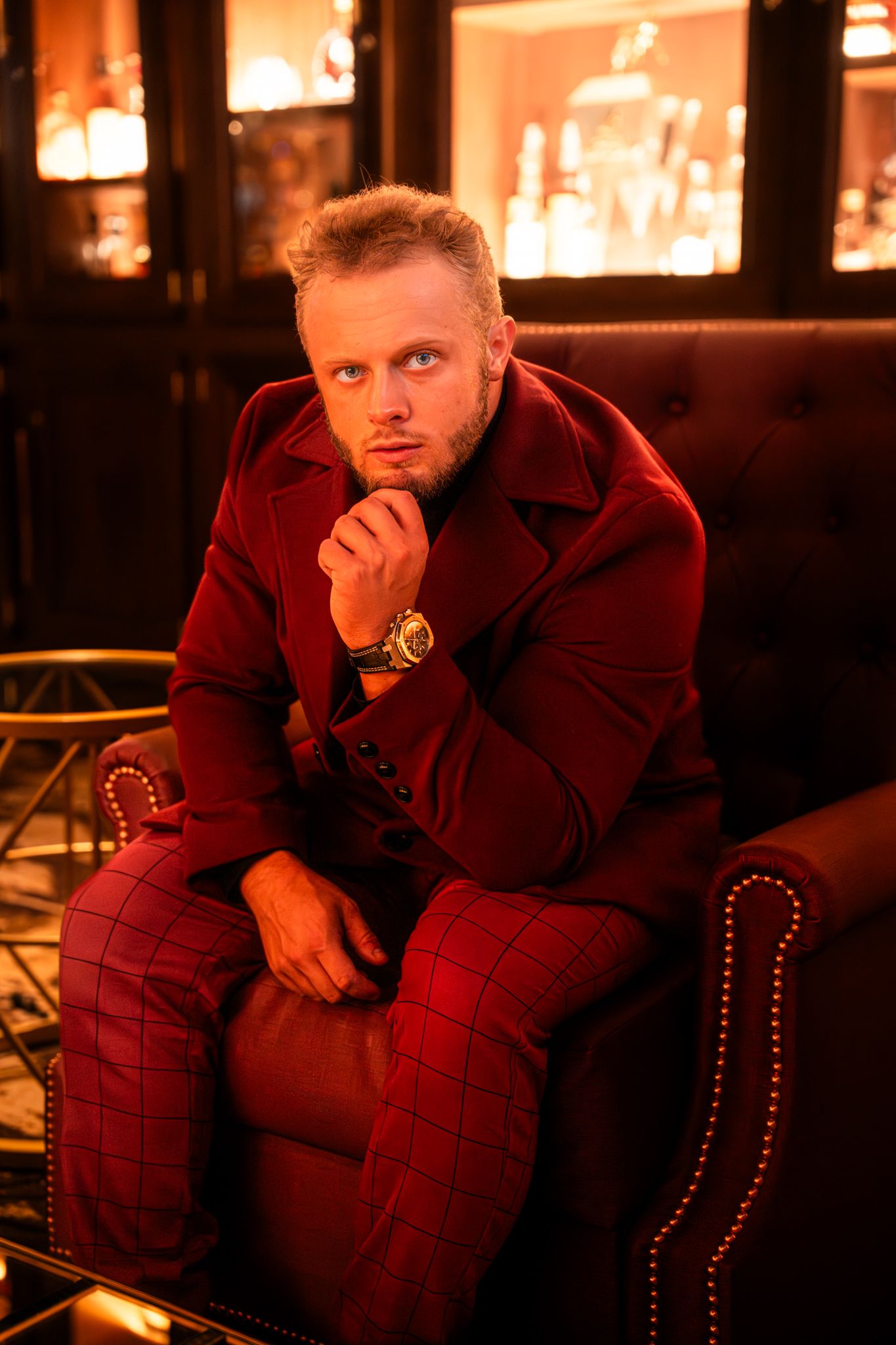 A person in a burgundy coat sits pensively in a dimly lit bar with warm ambient lighting and studded leather chairs.