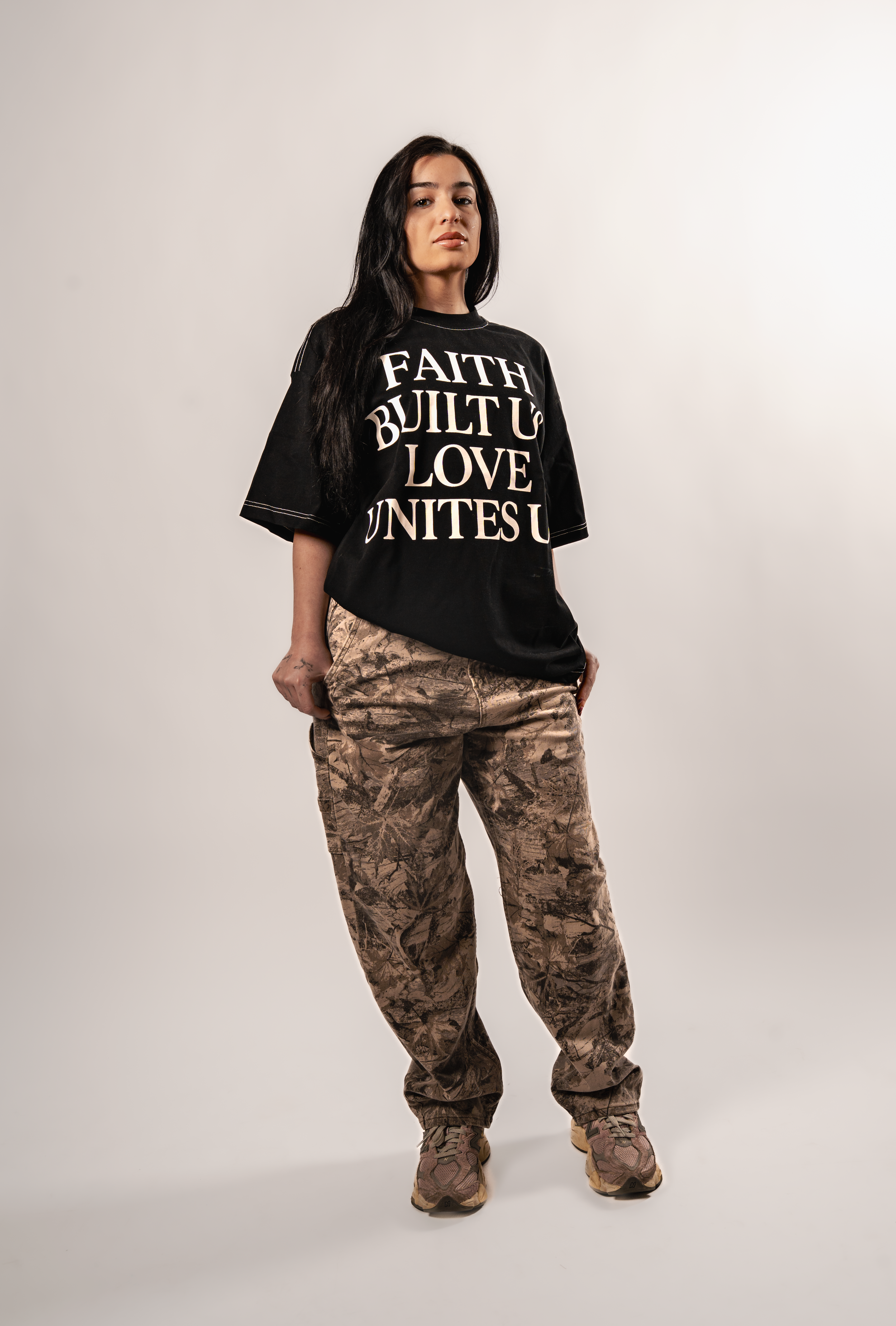 A person poses in a black graphic t-shirt and brown camouflage cargo pants against a white studio background.