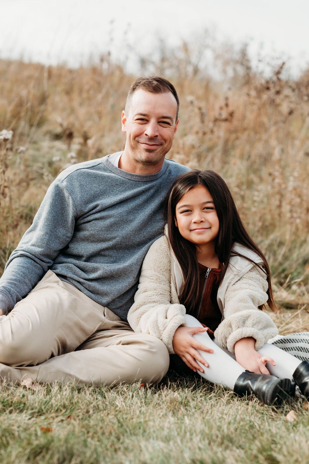 Relaxed couple laying in tall autumn grass during outdoor photoshoot wearing matching gray and beige outfits.