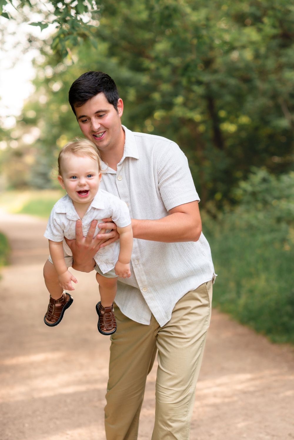 Father and baby playing together on a dirt path through lush green trees and bushes in a park setting.