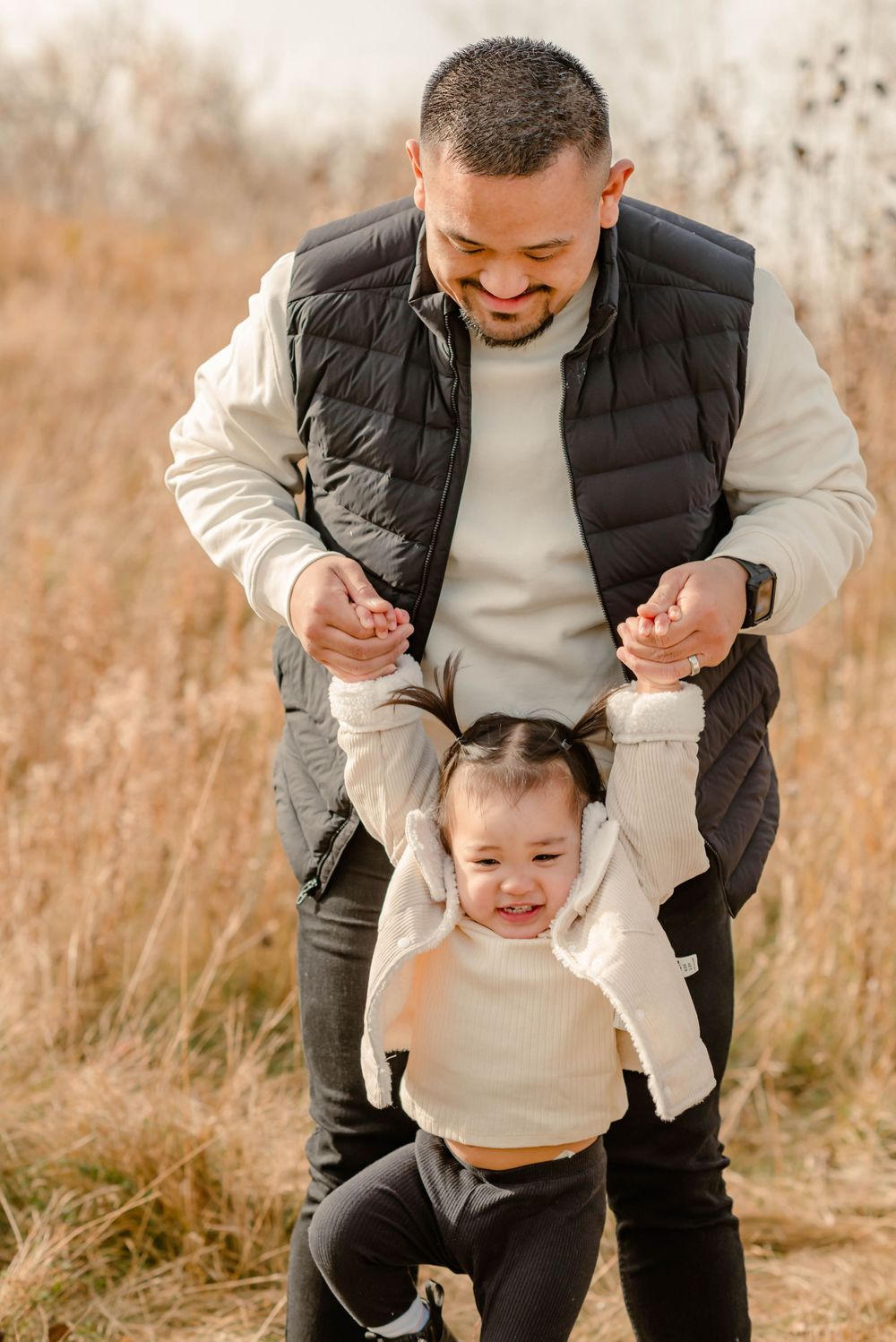 A parent and toddler play together in a field of tall brown autumn grass while wearing coordinated beige sweaters.