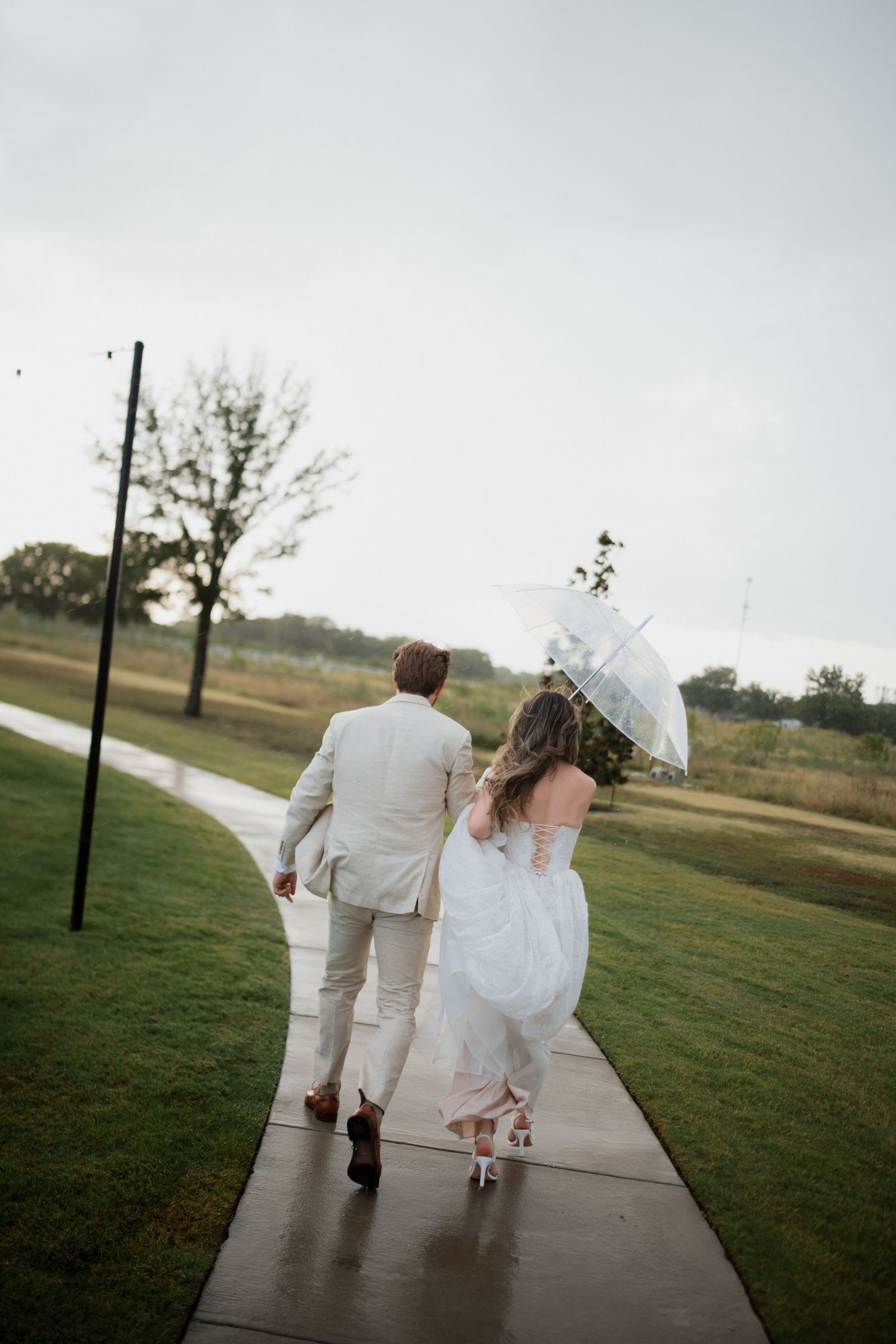 A couple in wedding attire walks down a path together holding a white umbrella on an overcast day.